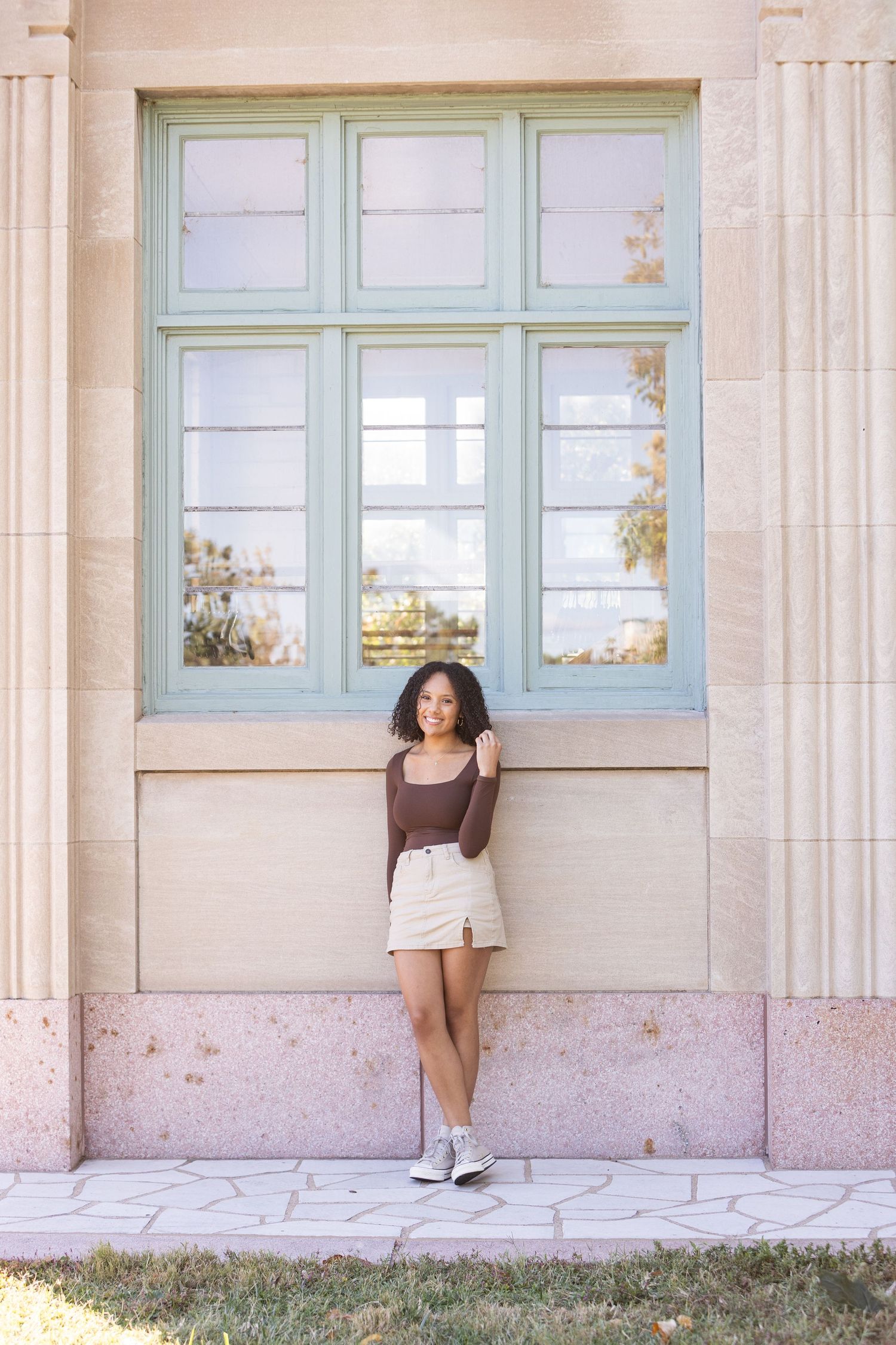 Young woman in brown top and white mini skirt posing against a pastel building with mint green windows.