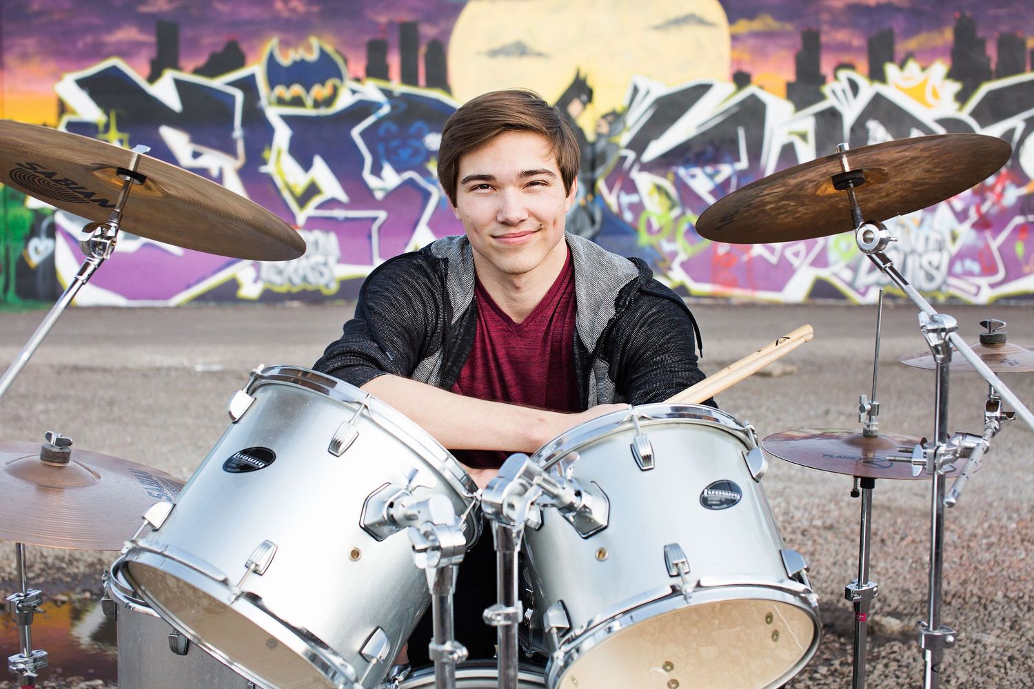 Young male drummer smiling behind a silver drum kit in front of colorful graffiti wall outdoors.