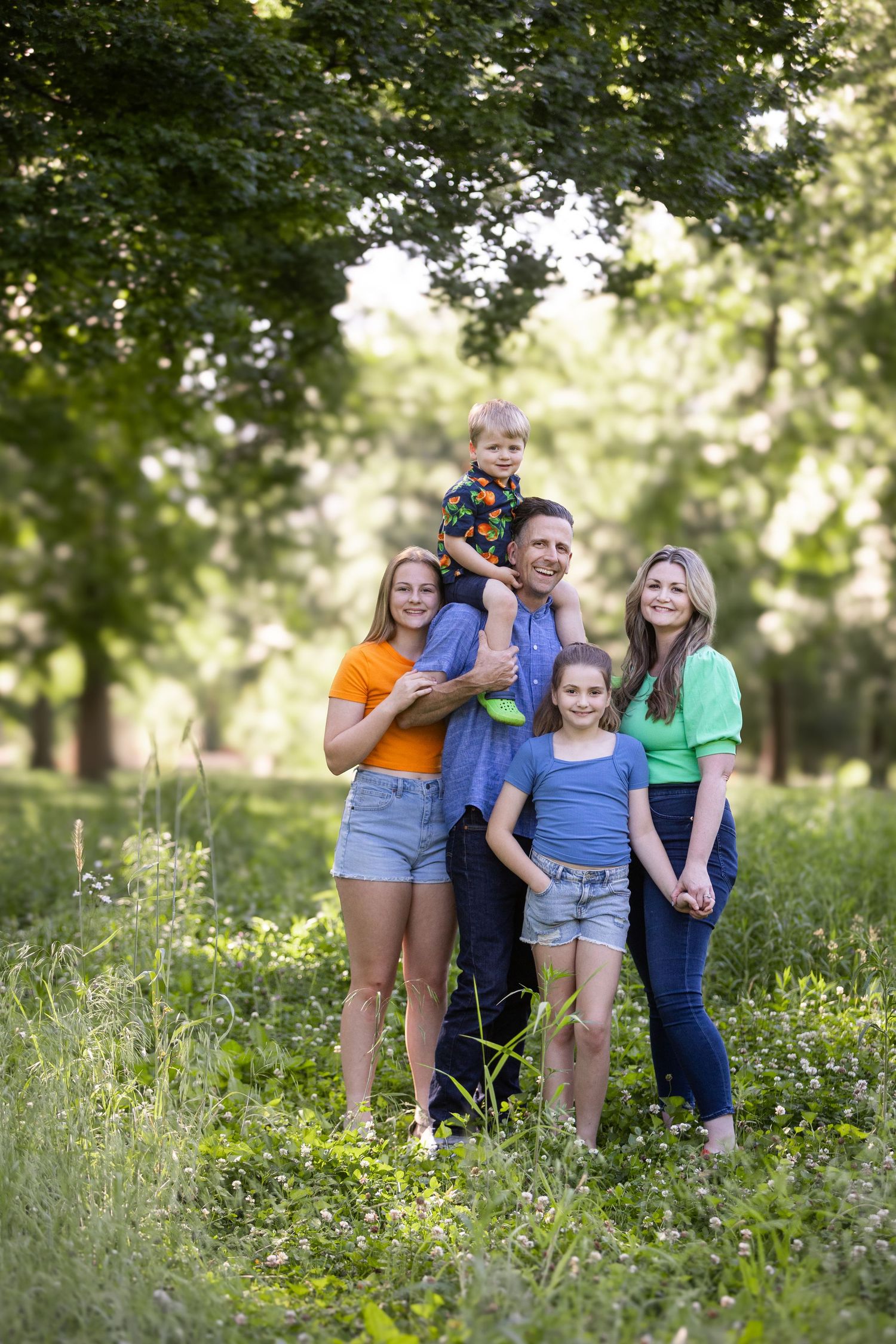 A happy family of six poses outdoors in a lush green park, with dad holding a toddler on his shoulders.