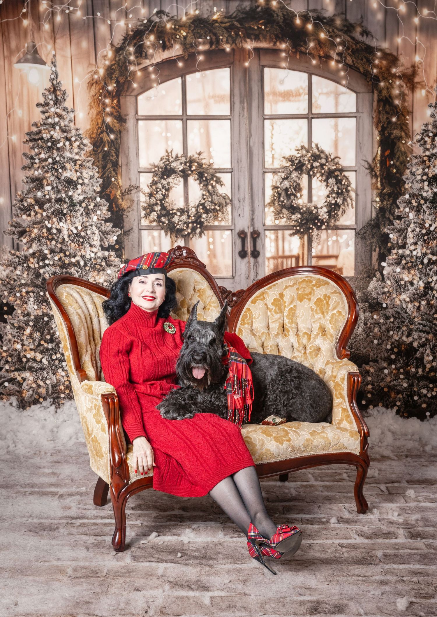 Woman in red dress and tartan hat sits with black dog on ornate sofa amid festive Christmas decor.