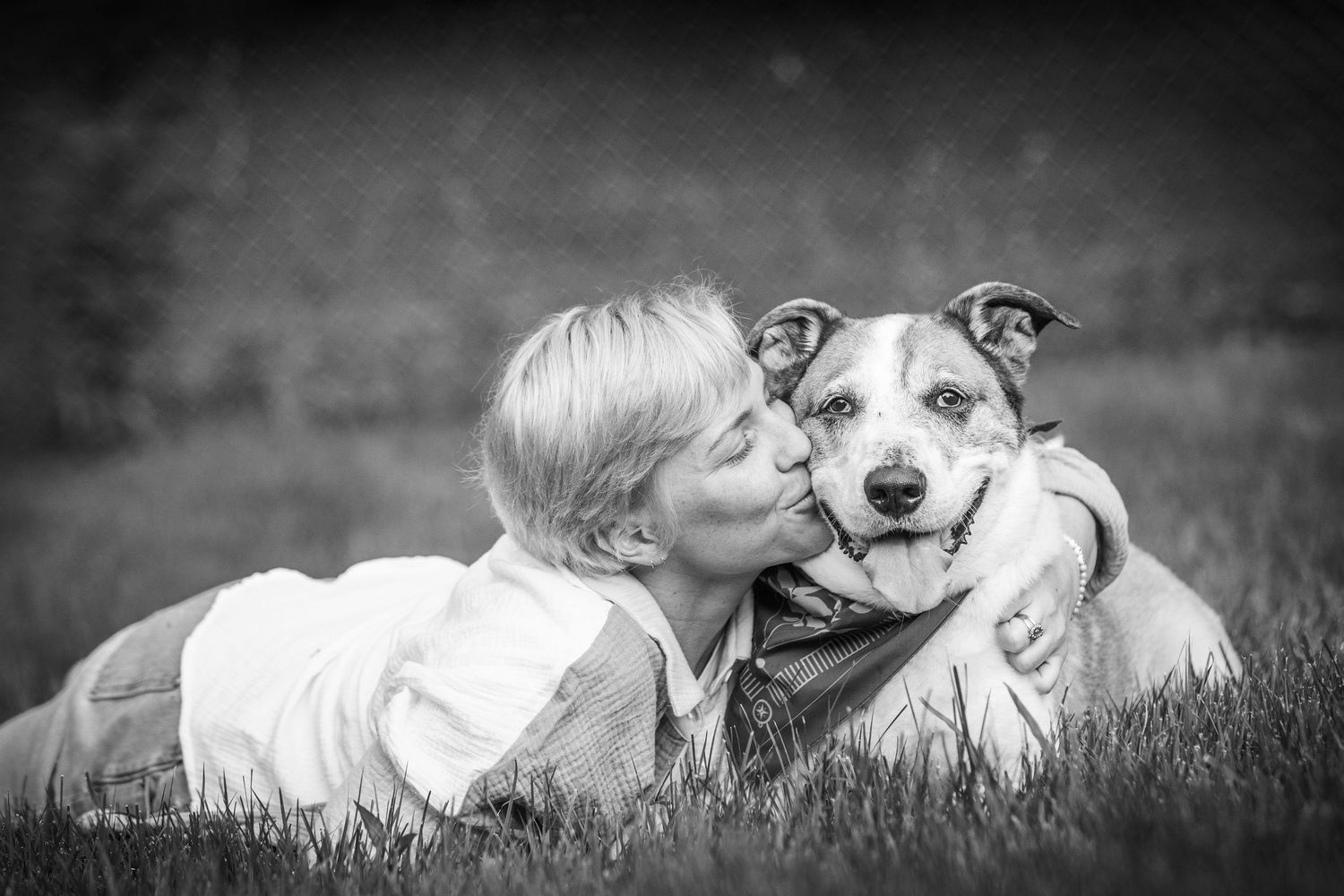 Black and white photo of a woman kissing her happy dog while lying in the grass outdoors.