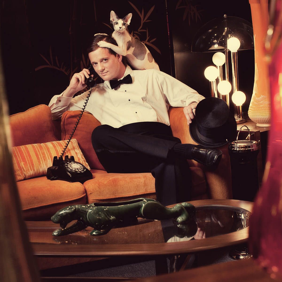 Retro photo of someone in formal wear lounging on an orange couch with a cat perched above.