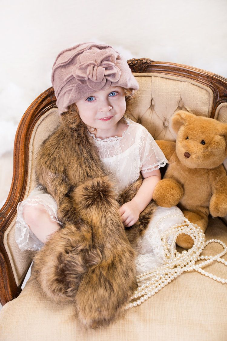 Child in vintage dress poses on chair with teddy bear and fur stole.
