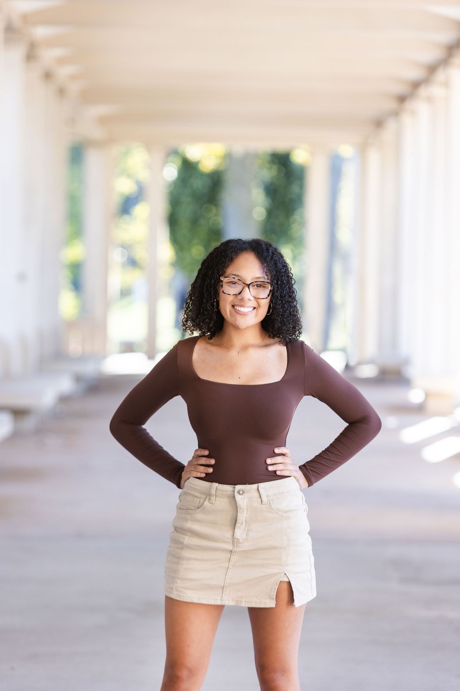 Young woman with curly hair and glasses wearing a brown top and beige skirt, posing confidently outdoors.