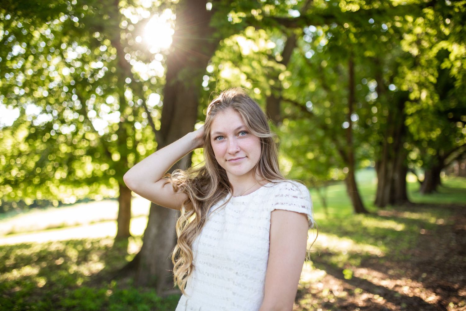 Young woman in white dress poses outdoors among tall trees with sunlight streaming through green leaves.