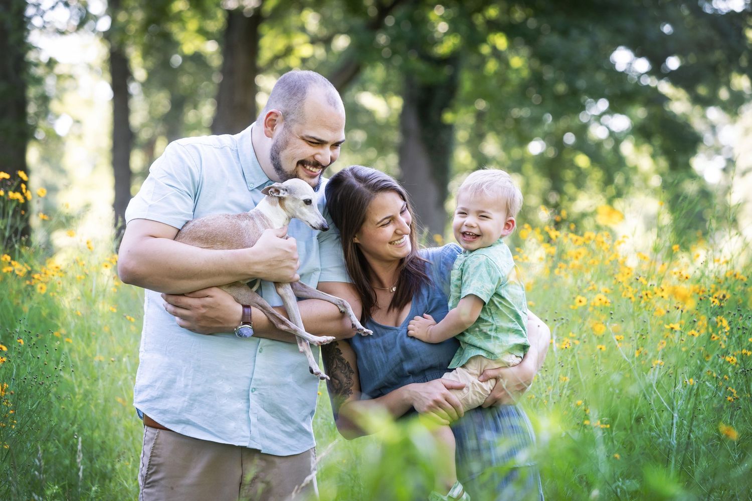 Happy family with a bald man, woman, toddler, and small dog posing in a sunny wildflower meadow.