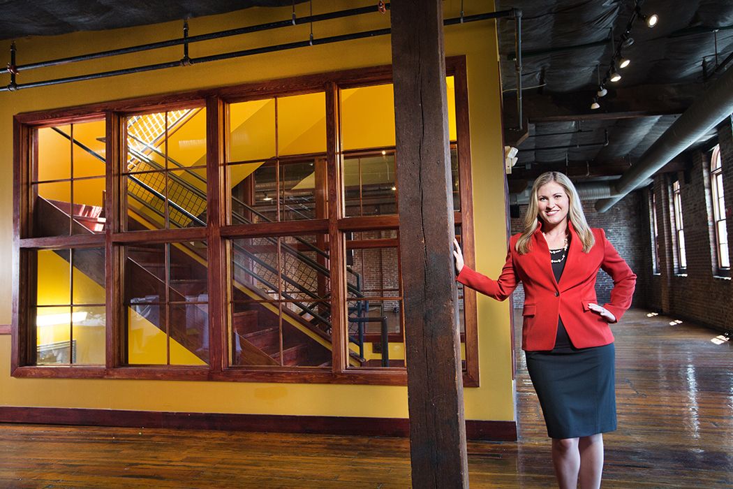 A professional in a red blazer stands in front of modern office windows with yellow accents.