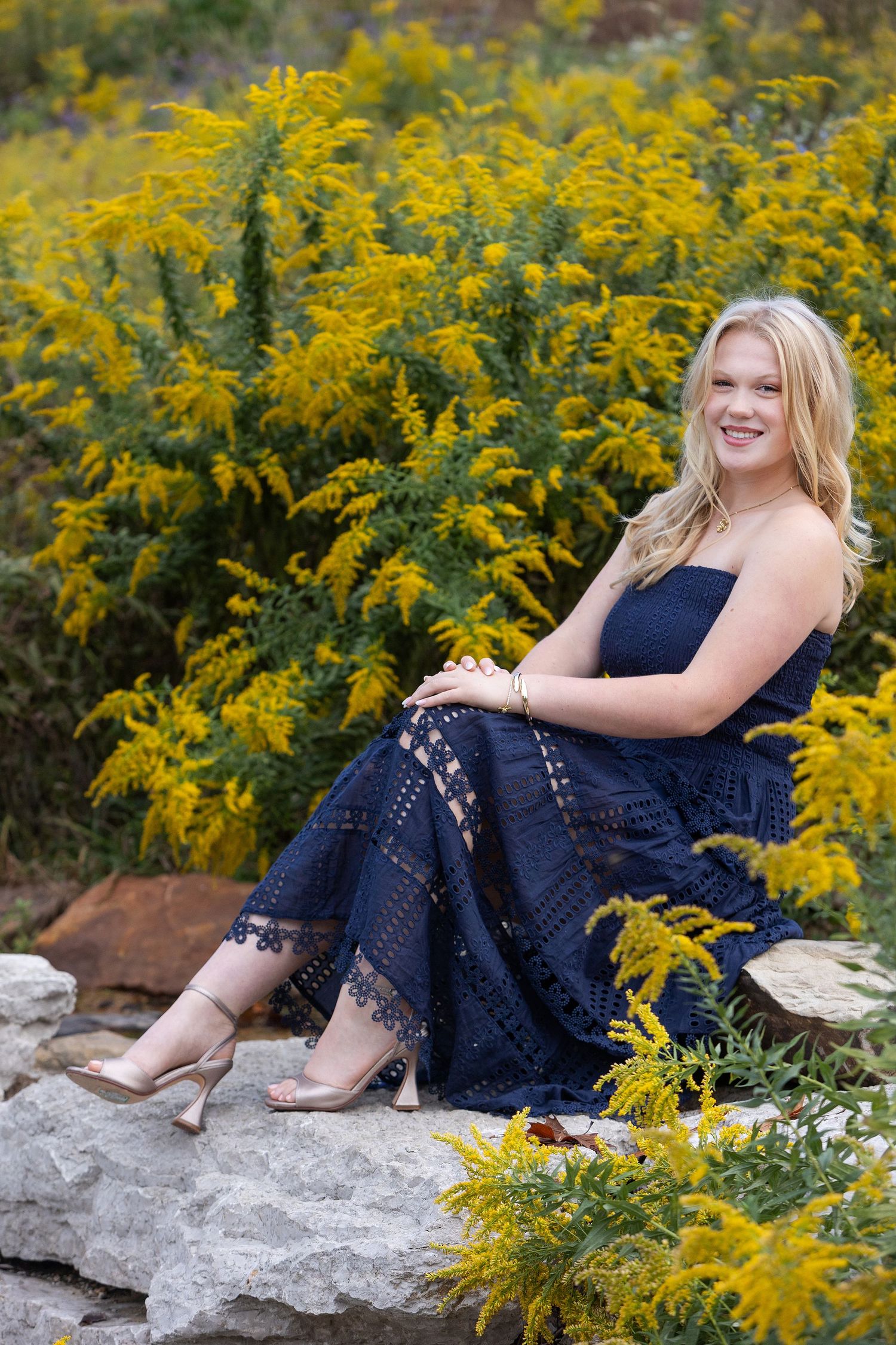 Young blonde woman in navy lace dress sitting on rocks surrounded by yellow wildflowers, smiling outdoors.