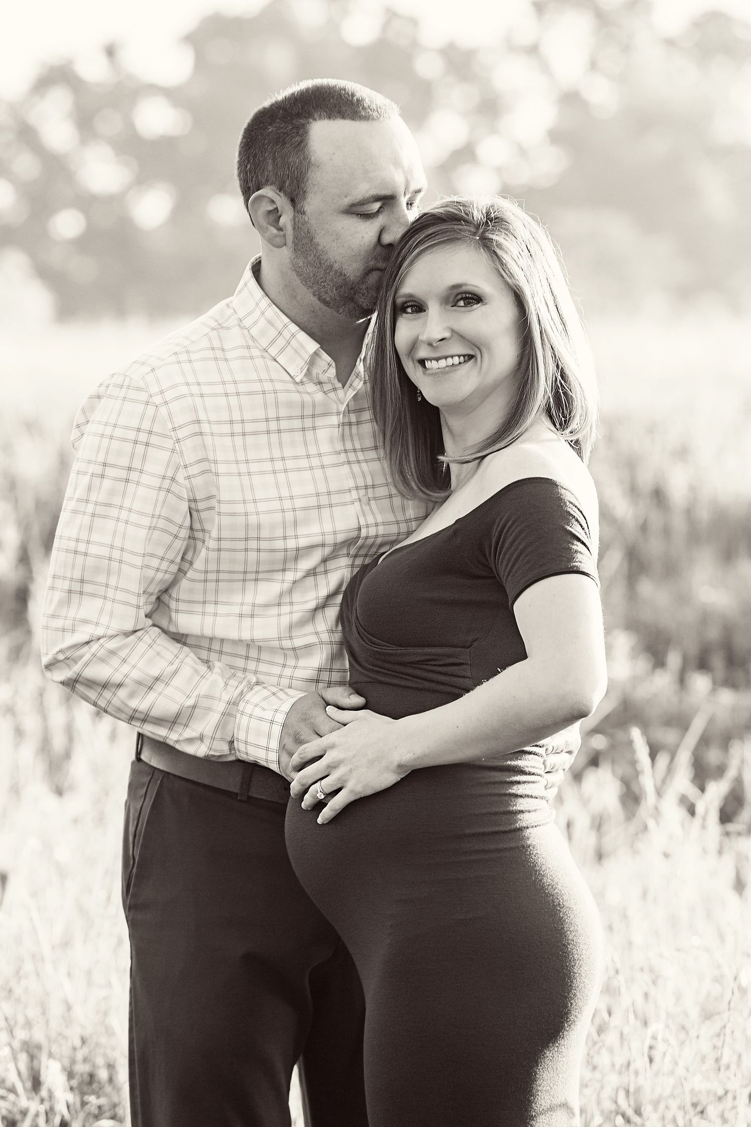 A loving couple embraces in a black and white maternity photo outdoors with soft bokeh lighting effects in the background.