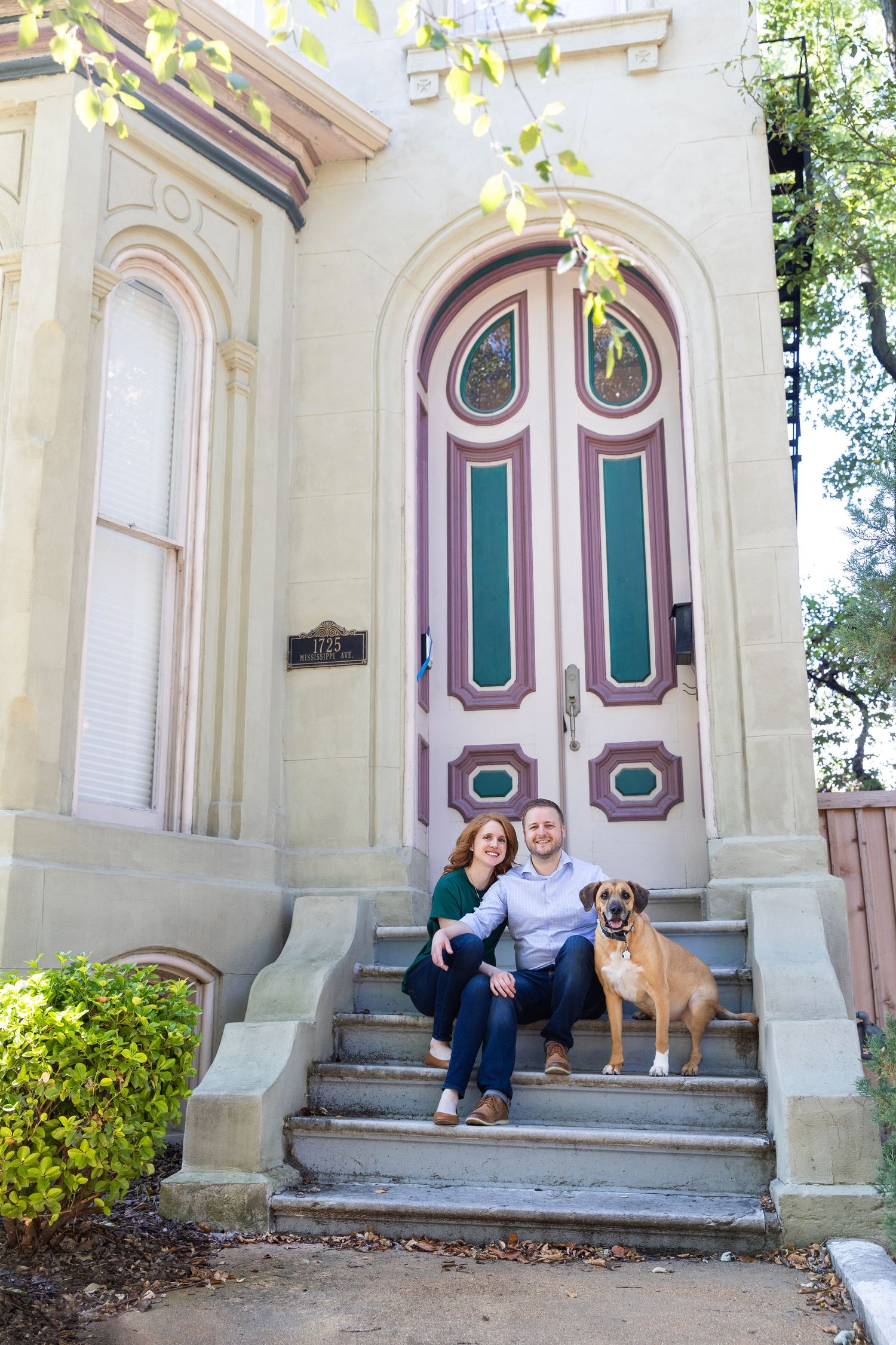 Family and dog sitting on steps of Victorian house with ornate teal and pink door.