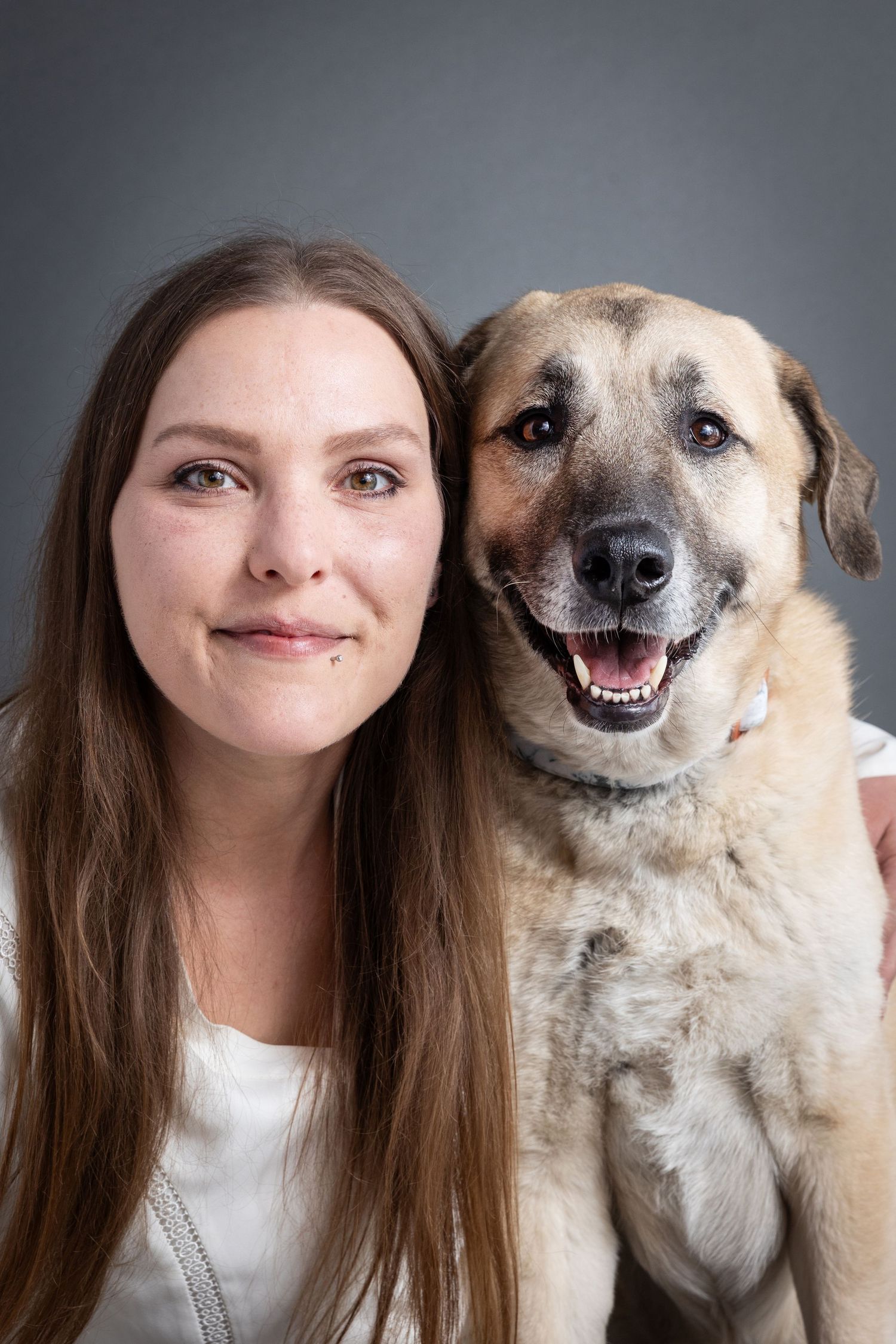 A smiling young woman with long brown hair poses with a happy tan dog against a gray studio background.