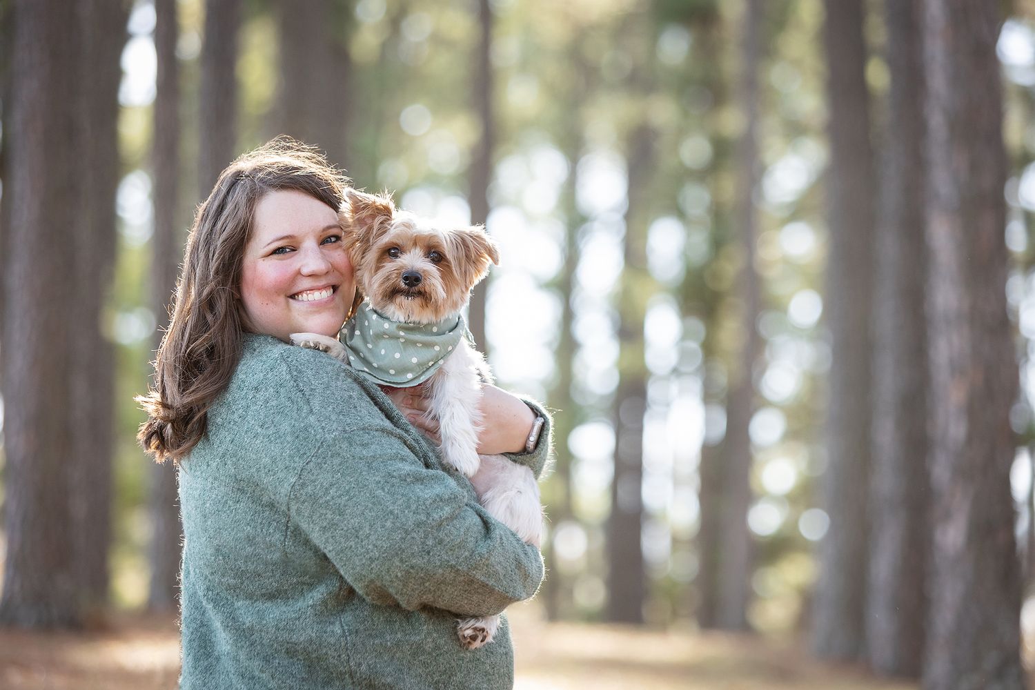 A smiling woman holds a small terrier dog while standing among tall pine trees in a forest setting.