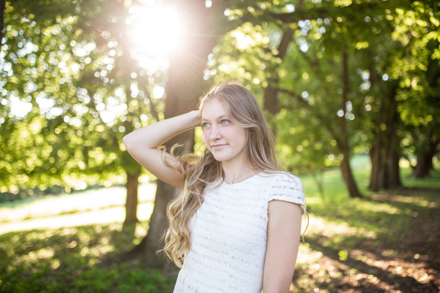 Young blonde woman in white lace dress poses in sunlit park with green trees glowing behind her.
