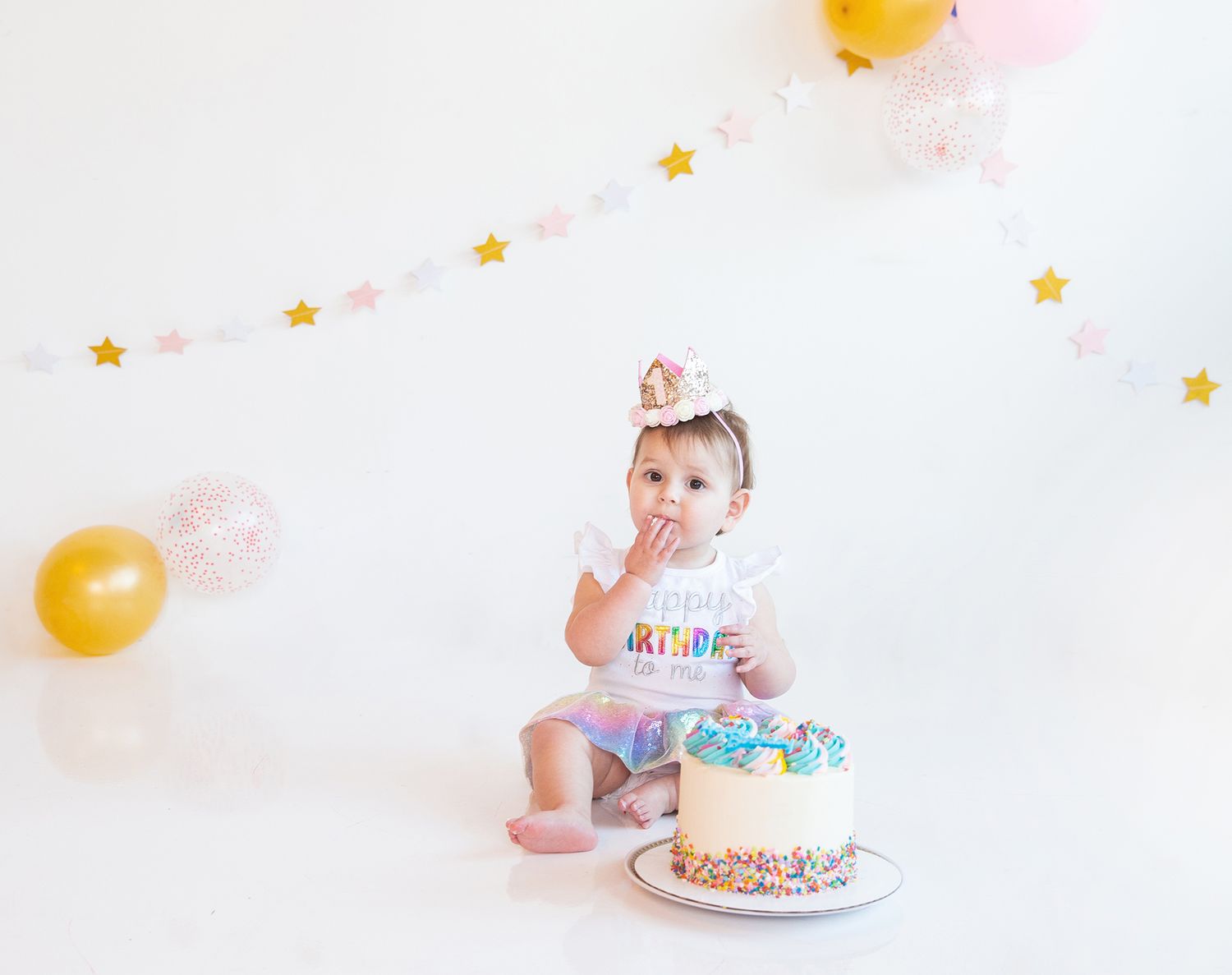 Baby's first birthday celebration with cake and balloons against white backdrop.