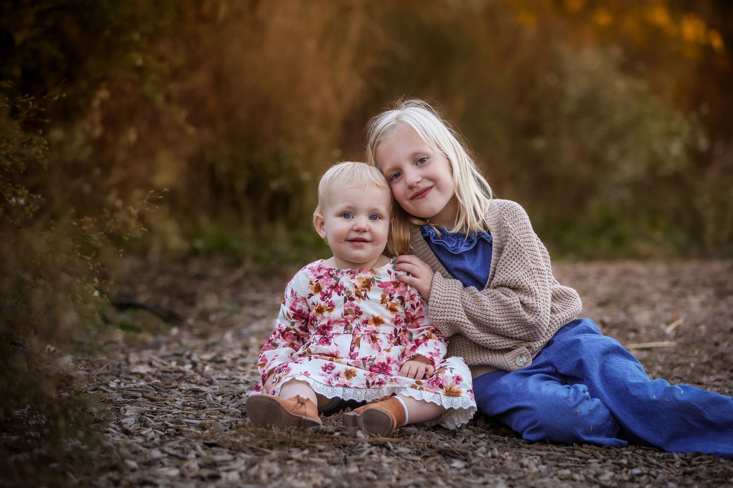 Two young sisters sitting together on a dirt path surrounded by autumn foliage, smiling warmly.