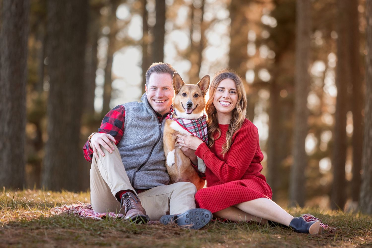 Smiling couple sitting outdoors with their Corgi dog wearing a bandana, surrounded by tall trees in autumn light.