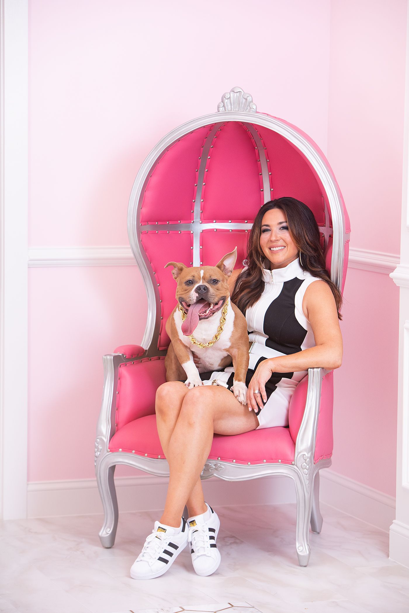 Woman in black and white outfit sitting in ornate pink chair with happy dog.
