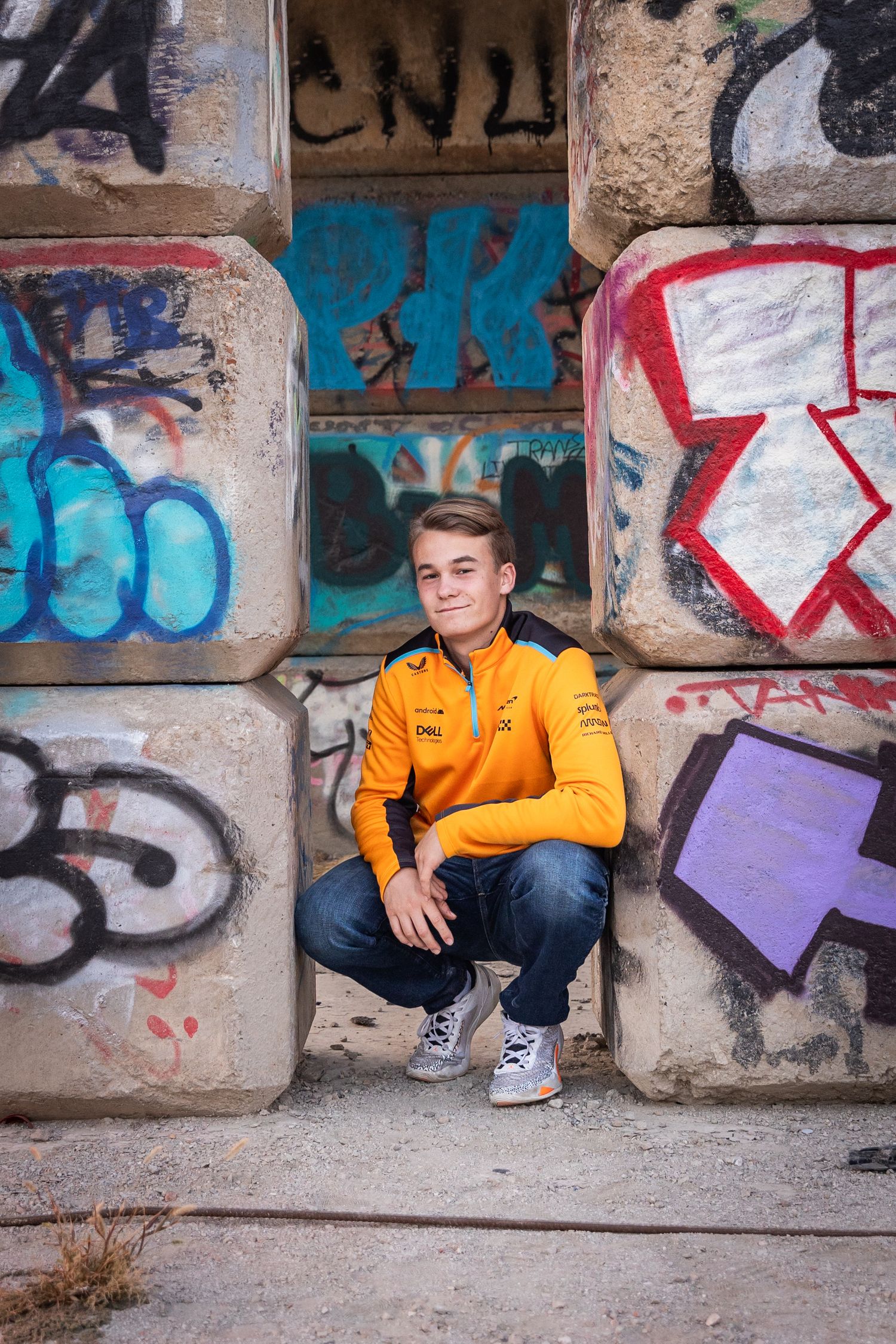 Young man in orange McLaren jacket crouching against graffiti-covered concrete blocks outdoors.