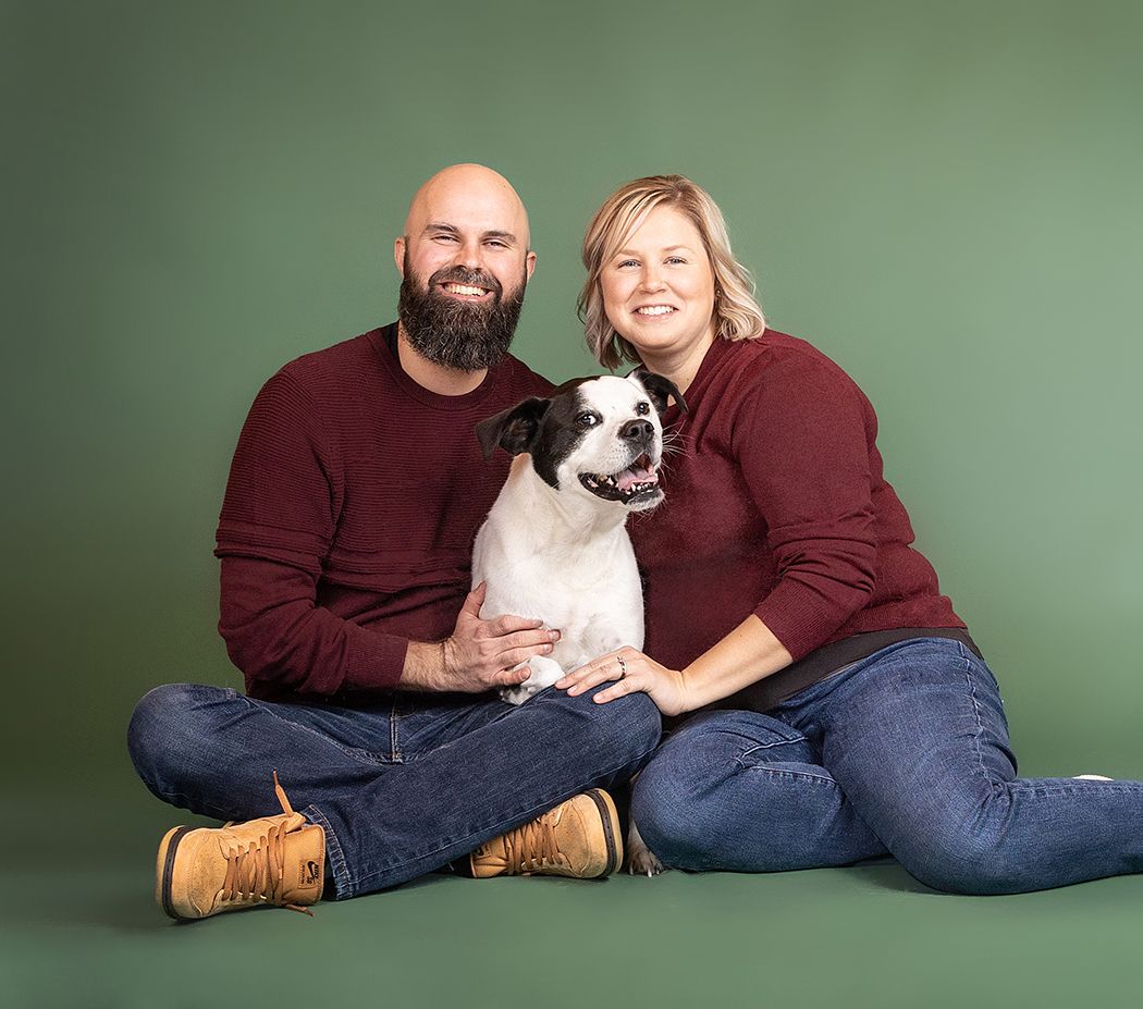 Portrait of couple with their black and white dog against green background.