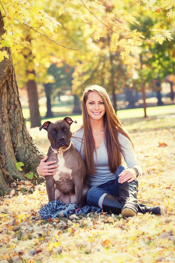 A dog owner and chocolate lab pose together among fallen autumn leaves in a sunlit park.