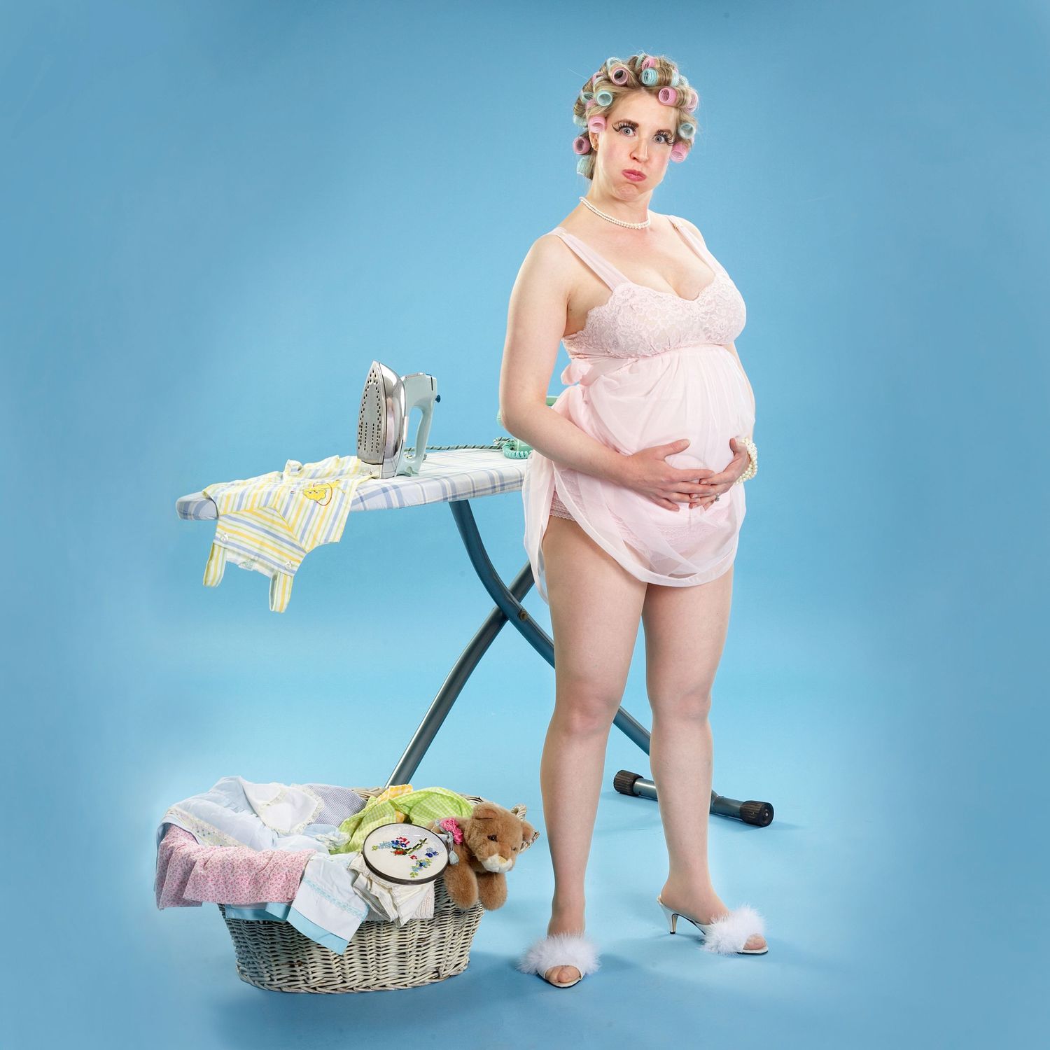 Vintage-style portrait in pink undergarments with laundry basket and ironing board against blue background.