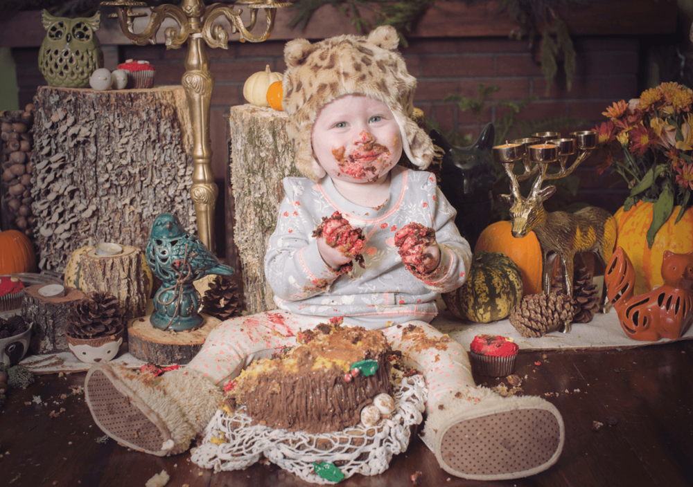Baby in white outfit surrounded by pumpkins and autumn decor making messy face while eating.