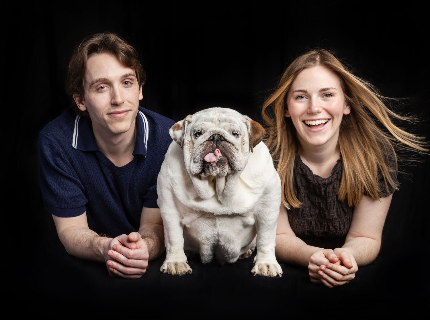 Two smiling young adults posing with a white English Bulldog against a black background.