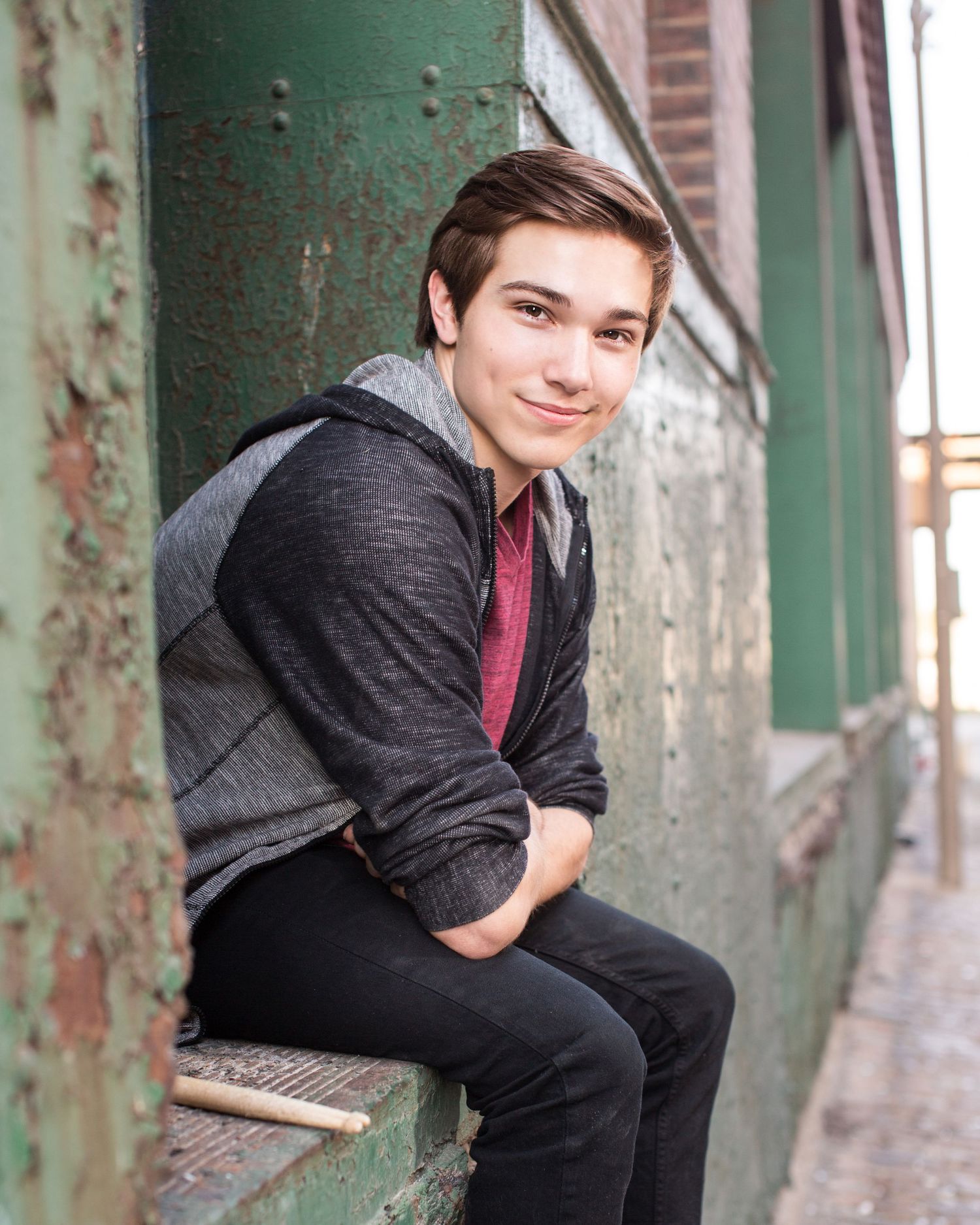 A young man in a dark hoodie and red shirt sits relaxed against a weathered green wall in an urban alley.