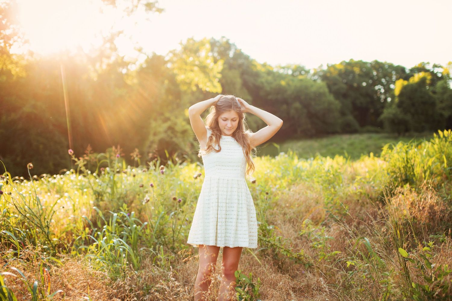 Young woman in white dress and cowboy boots standing in a sunlit field with golden hour light streaming through trees.