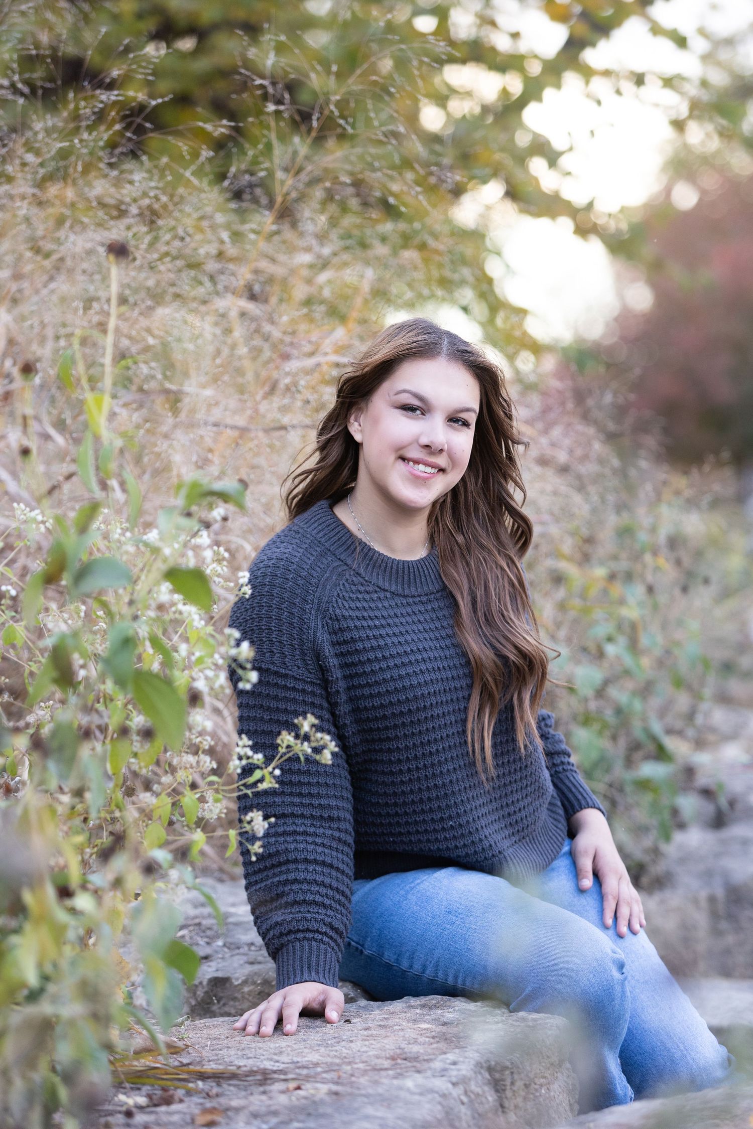 Young woman with long brown hair smiling outdoors, wearing a gray knit sweater and jeans, seated on rocks among autumn foliage.