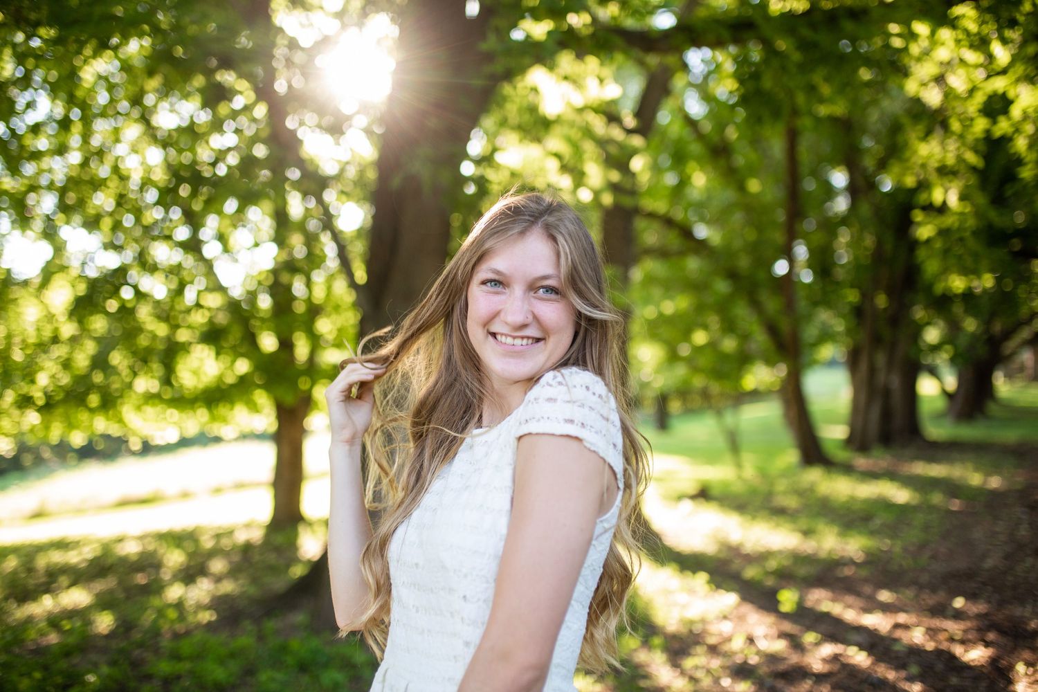 Smiling young woman with long blonde hair in a white dress standing among sunlit green trees in a park.