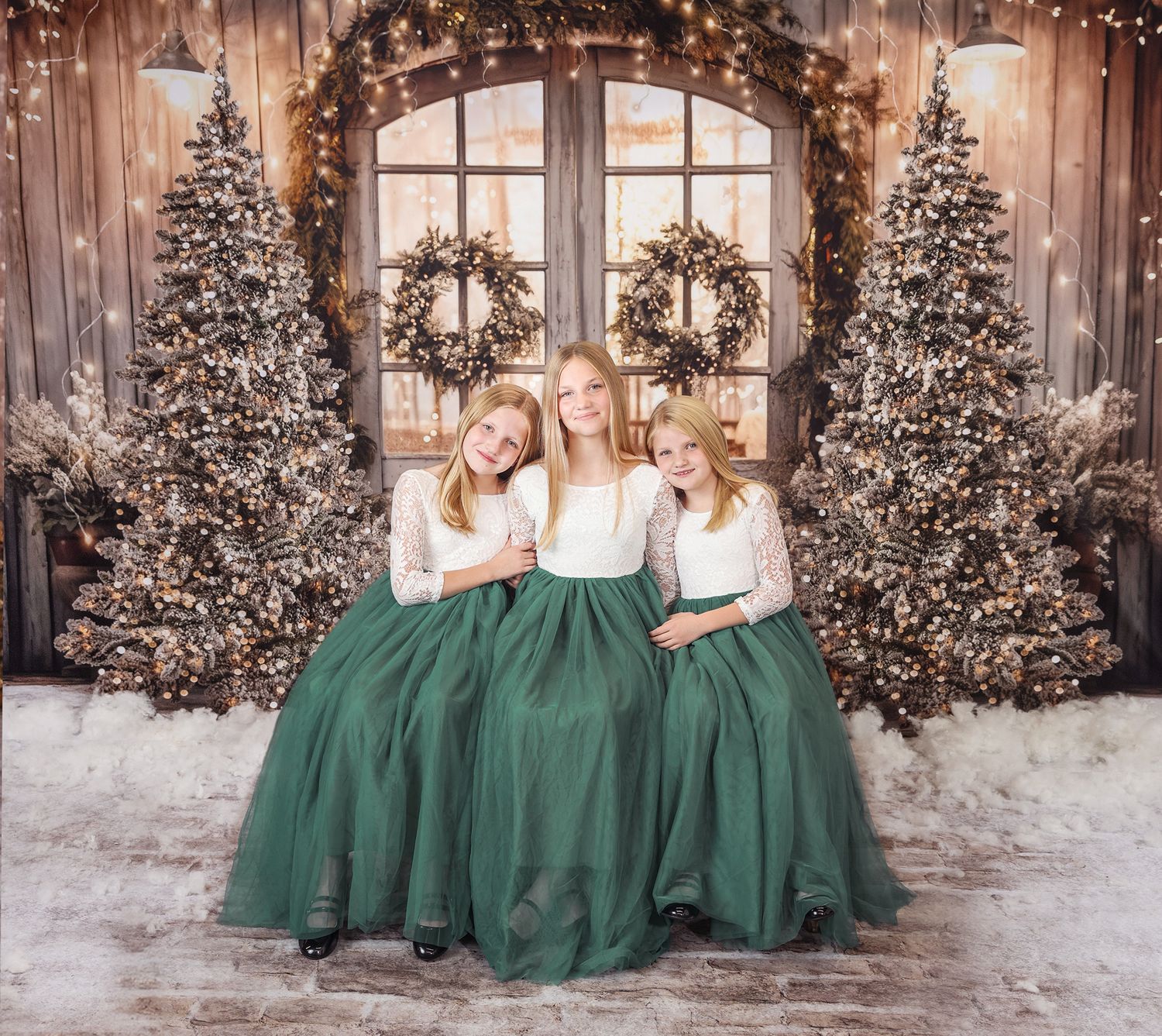Three people in matching white tops and emerald green tulle skirts pose in front of decorated Christmas trees and wreaths.