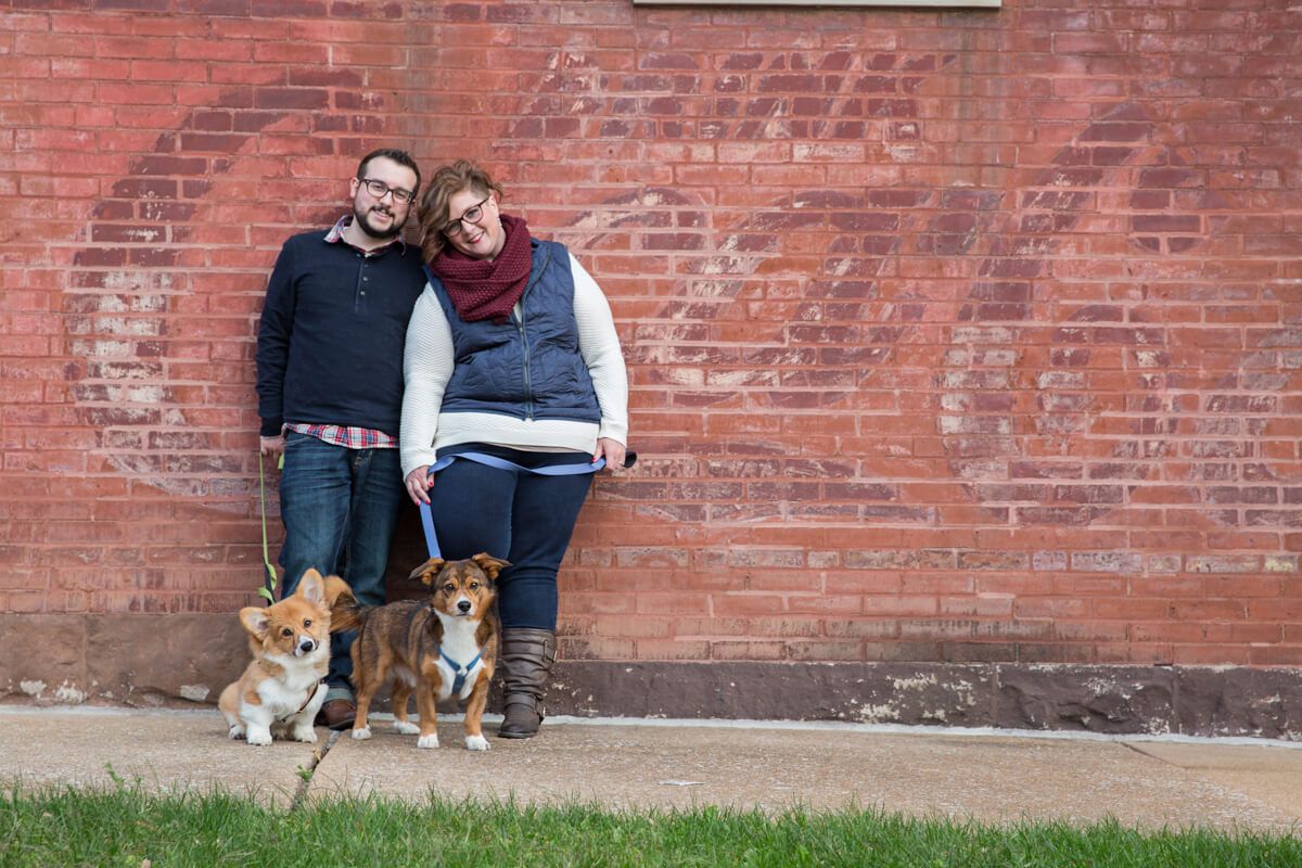 Couple leans against brick wall with their two Corgi dogs in casual outdoor portrait.
