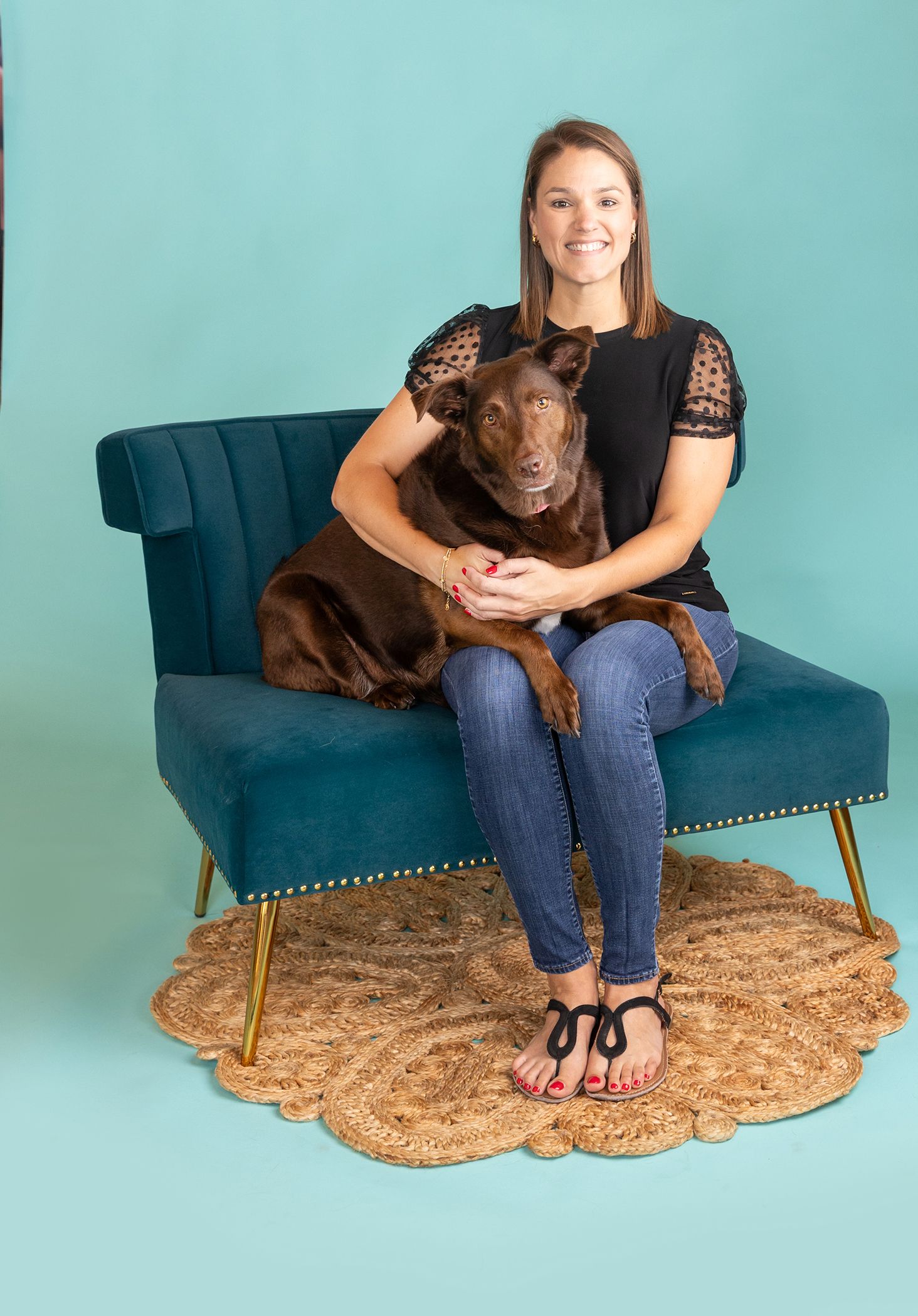 Woman smiling on teal velvet chair with chocolate Labrador dog, teal background, jute rug, studio portrait.