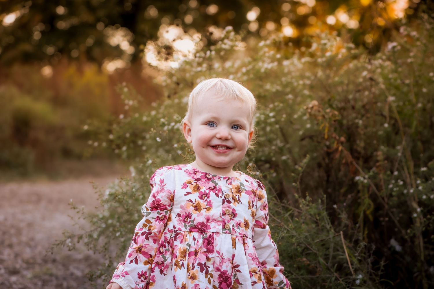 Smiling toddler girl in floral dress standing outdoors among wildflowers in golden hour light.