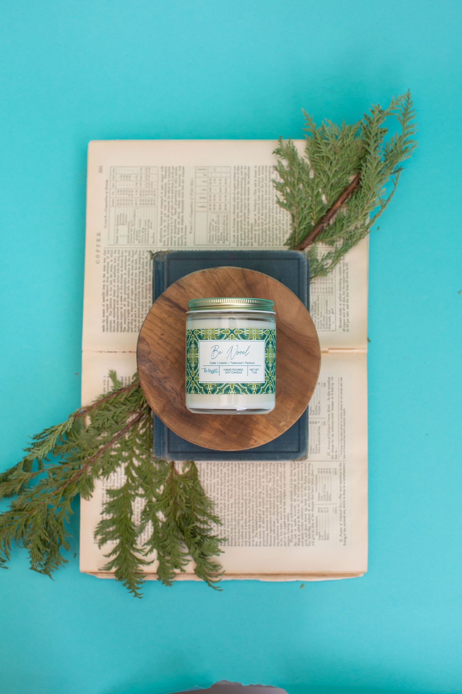 Vintage book displays candle jar with cedar sprigs on turquoise surface.