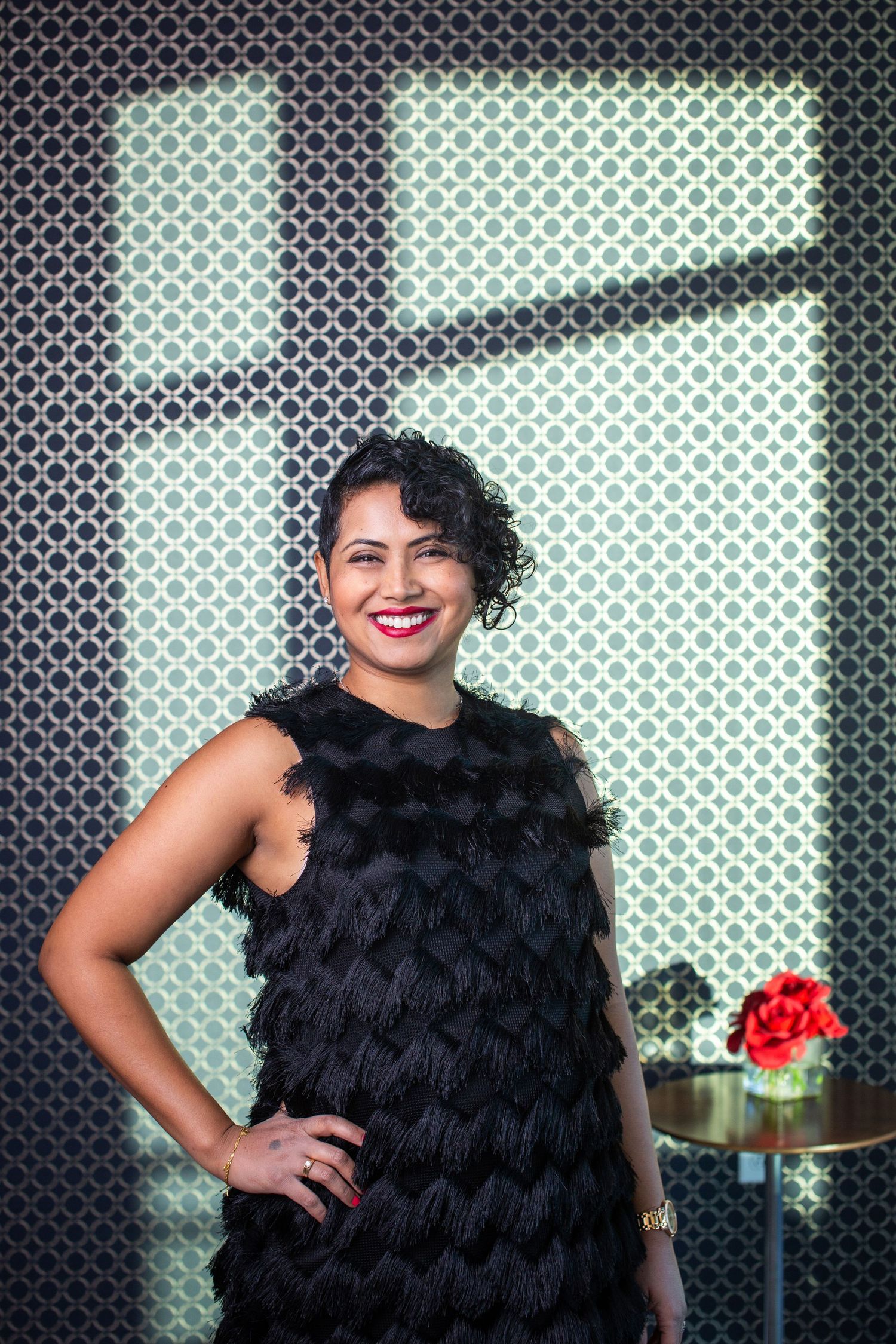 A person wearing a black feathered dress poses confidently against a geometric patterned background.