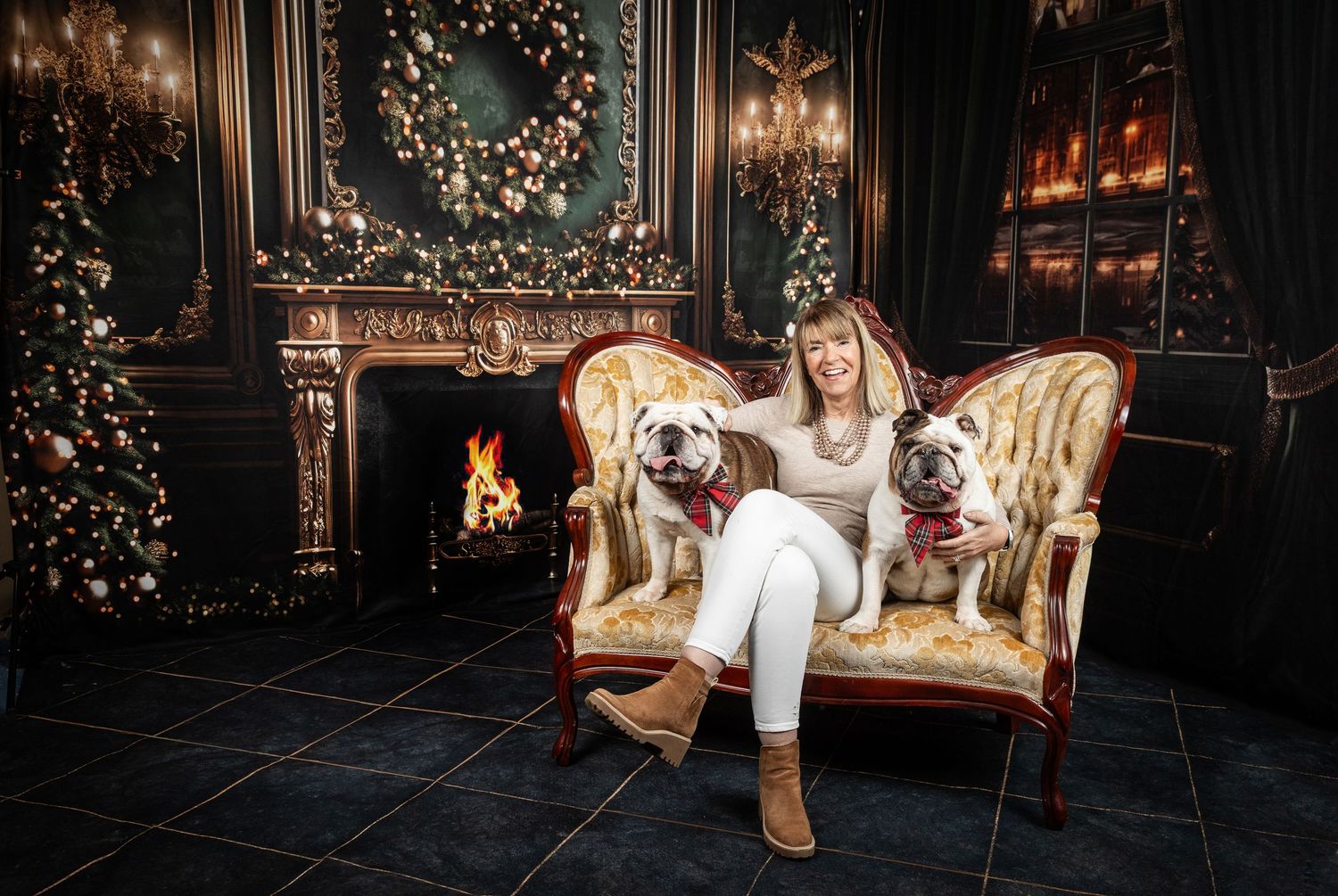 Woman sitting on ornate chair with two white bulldogs beside a fireplace and Christmas tree in elegant holiday decor.