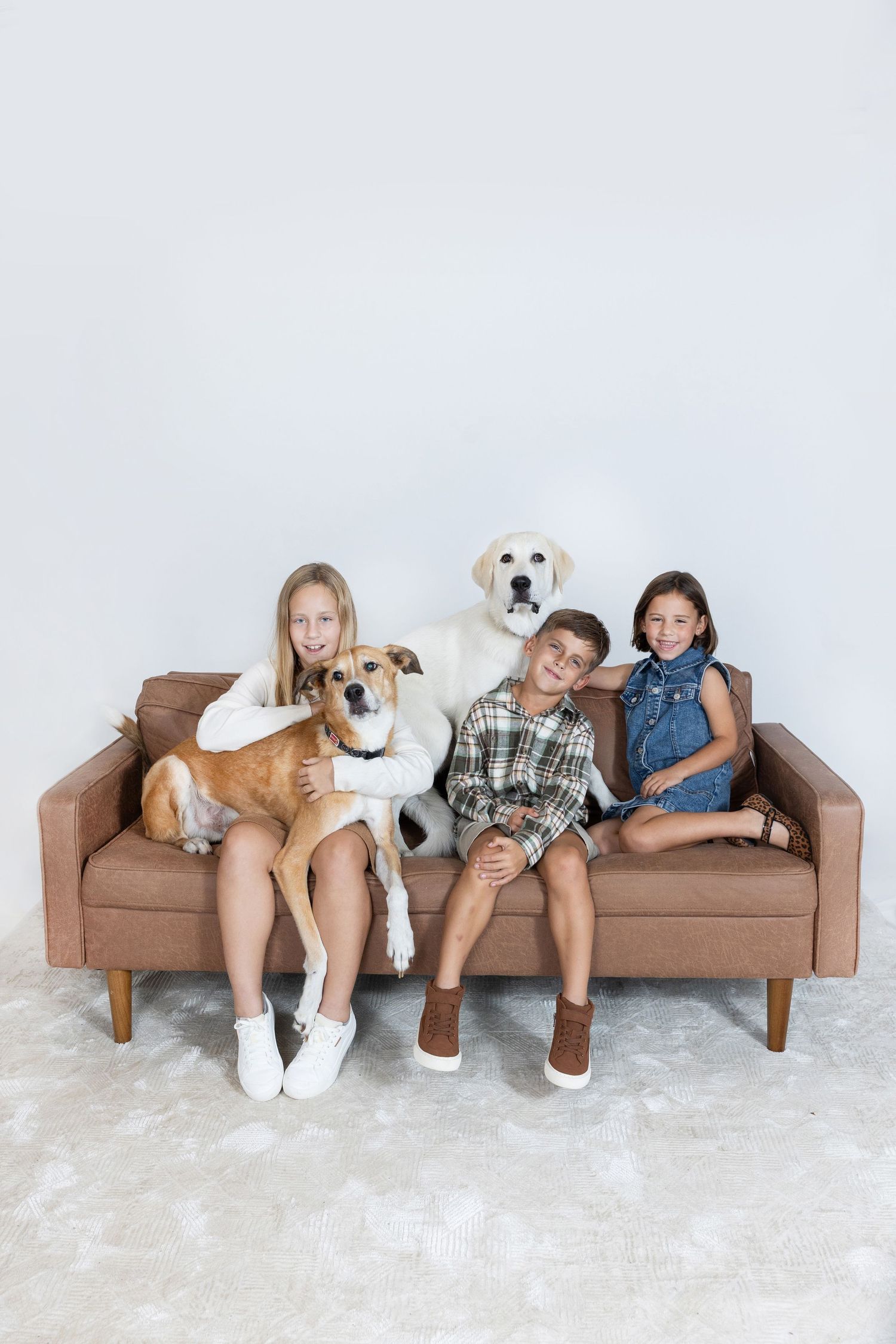 Three children sitting on a brown sofa with two dogs, a golden Labrador and a tan mixed breed, against a white wall.