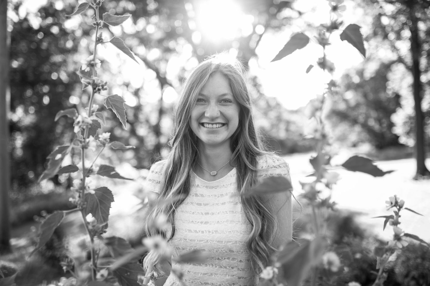 Smiling young woman with long blonde hair posing outdoors among lush green leaves in a black and white photo.