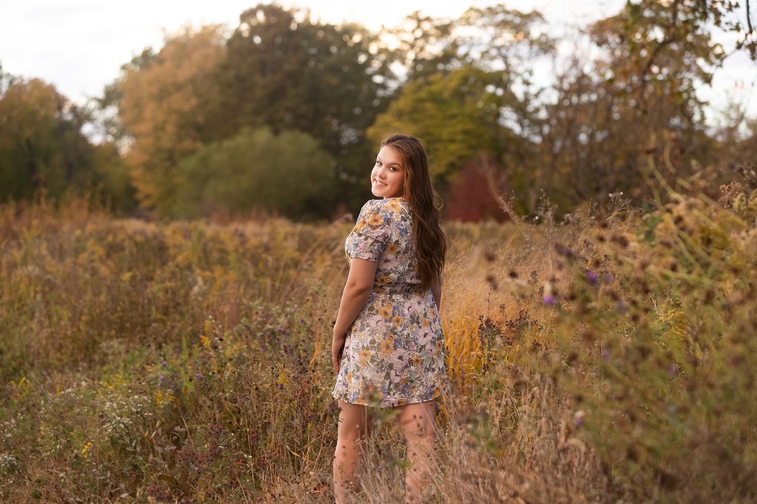 Young woman in floral dress standing in golden autumn field with trees in background during sunset.