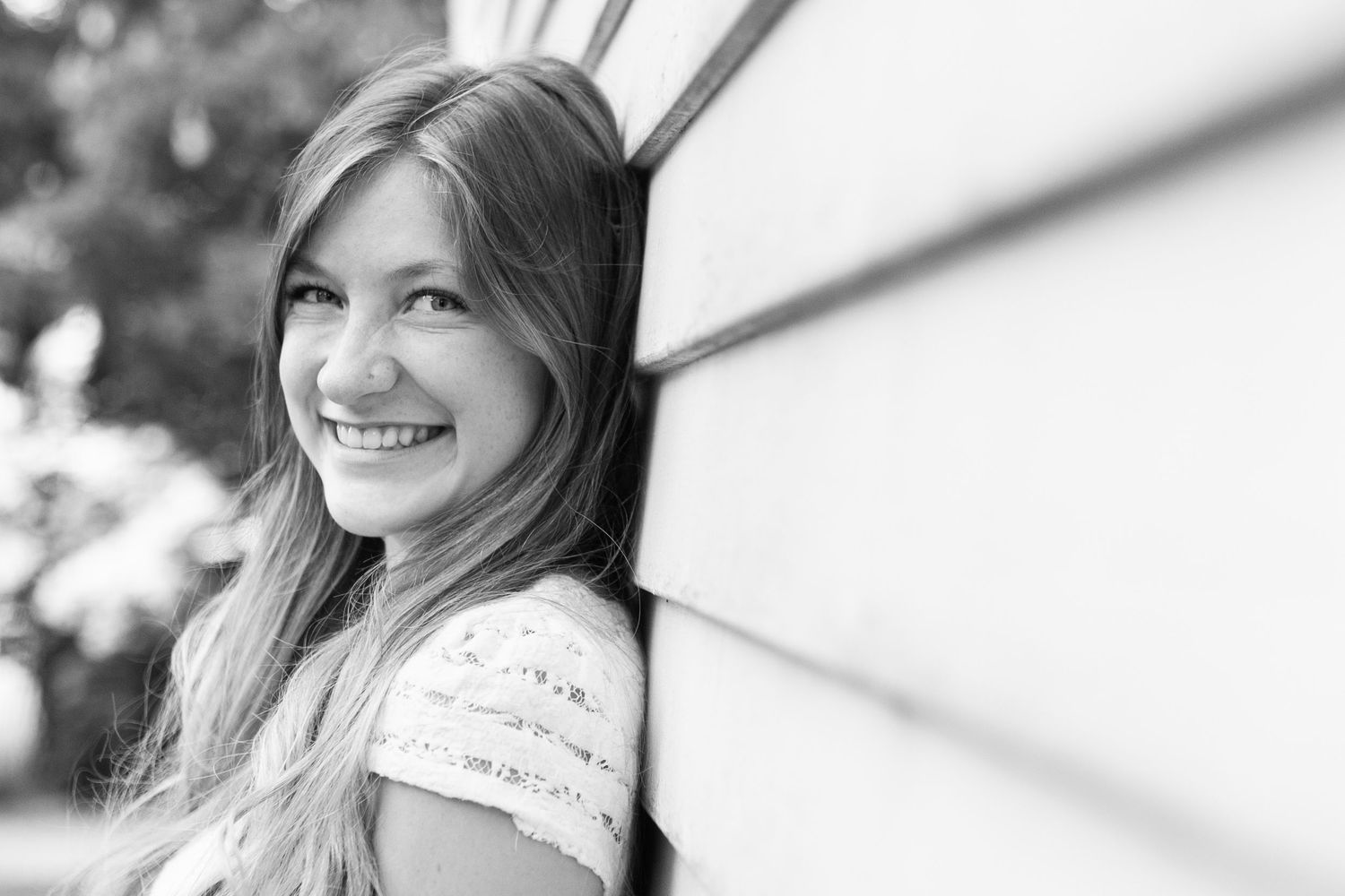 Smiling young girl with long hair leaning against a wall outdoors in a black and white portrait photo.