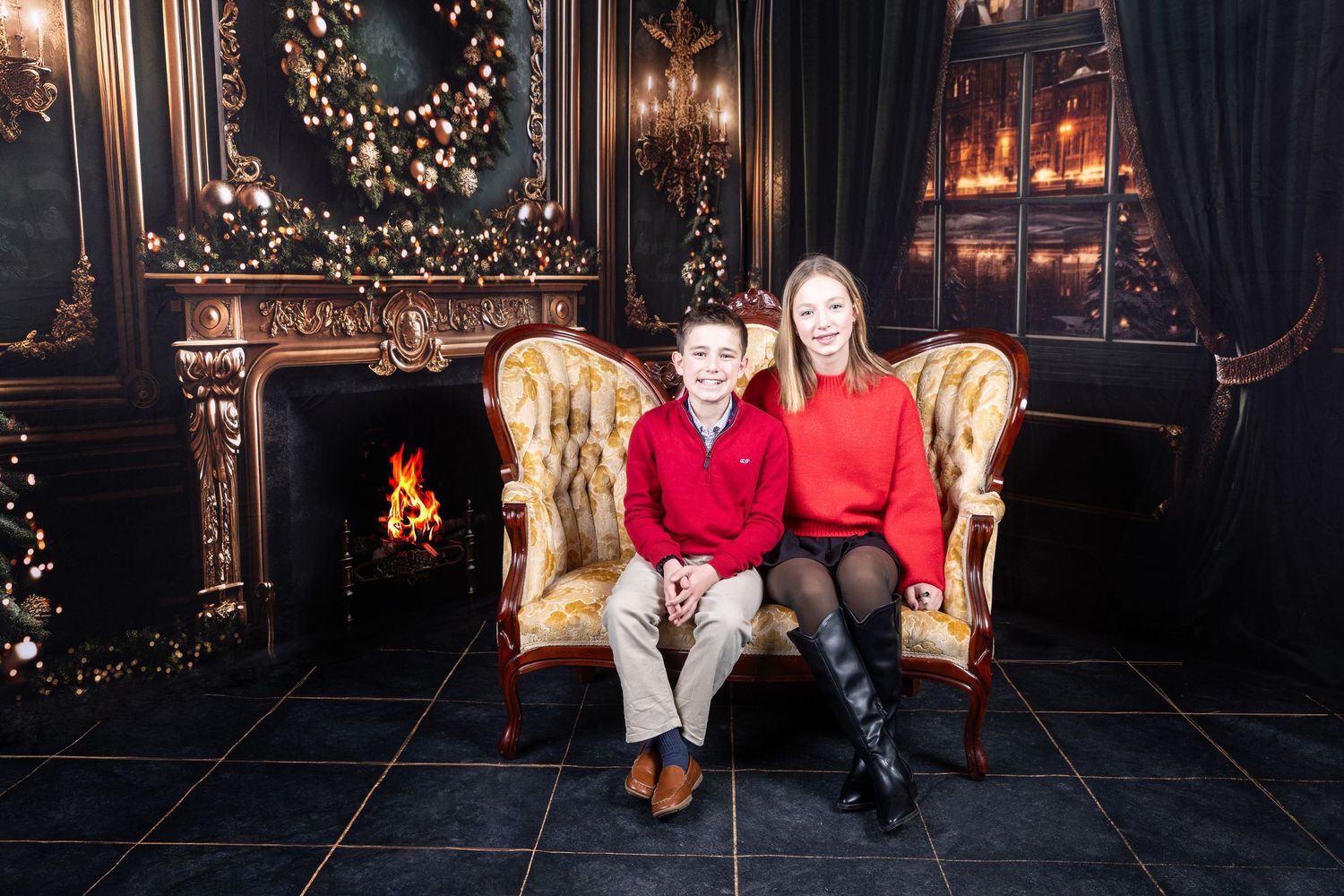 Two children in red sweaters sit on a gold chair by a fireplace with Christmas decorations in a festive holiday portrait.