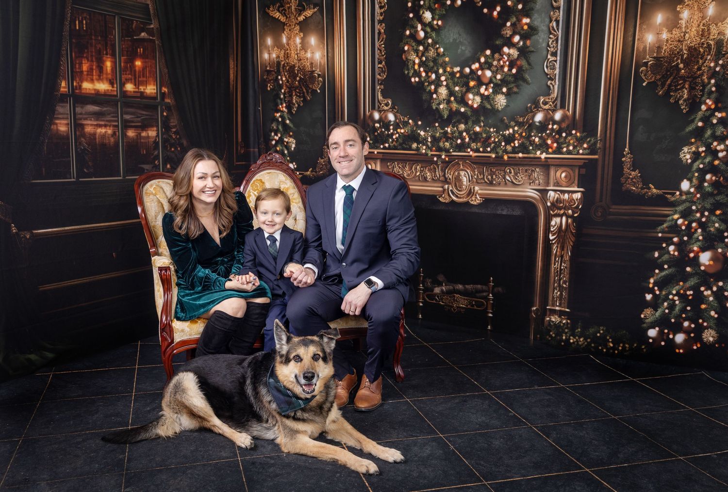 Family Christmas portrait with parents, young boy, and German Shepherd dog posed by decorated fireplace and holiday tree.