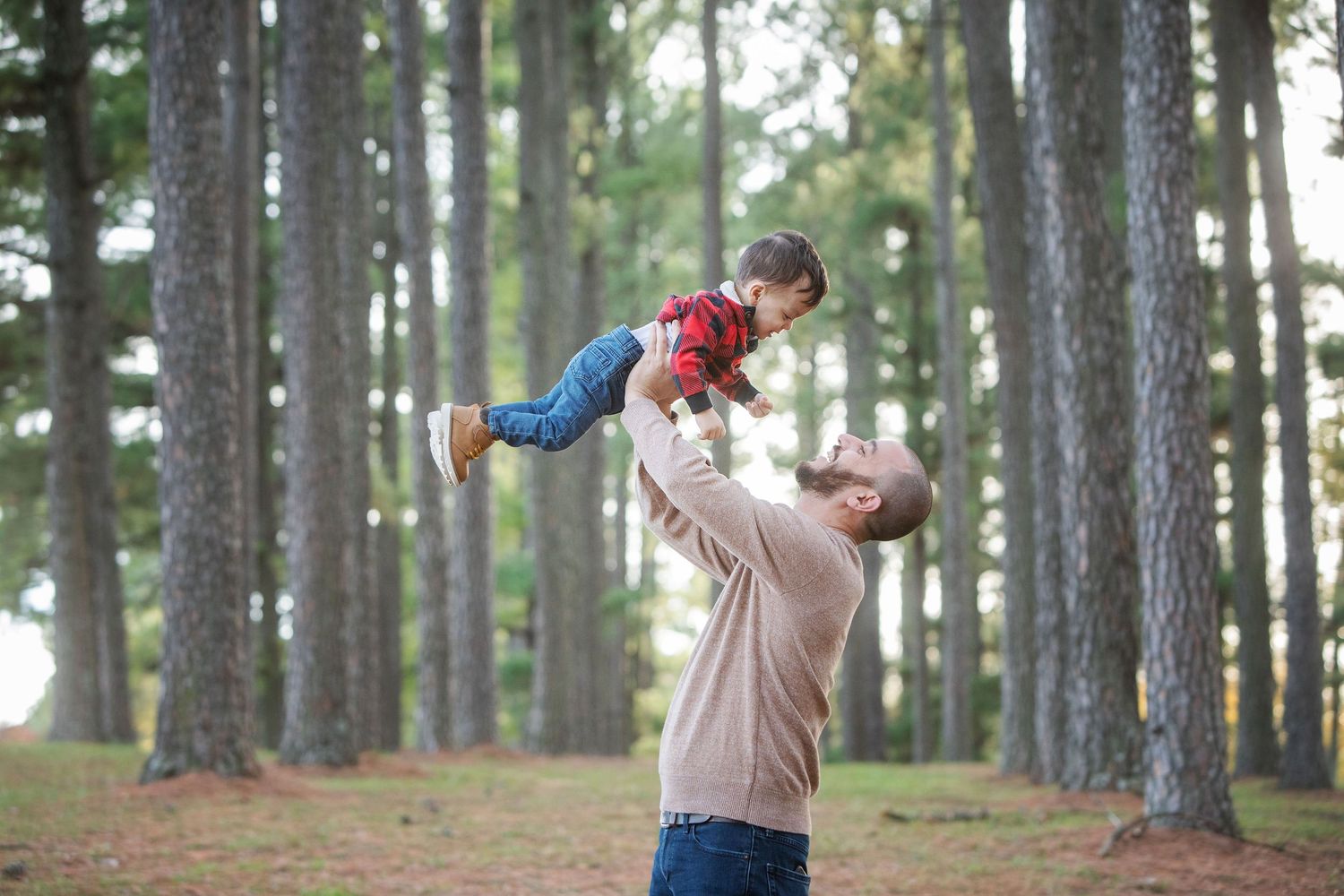 Father joyfully lifting his young child in the air outdoors among tall pine trees in a forest setting.