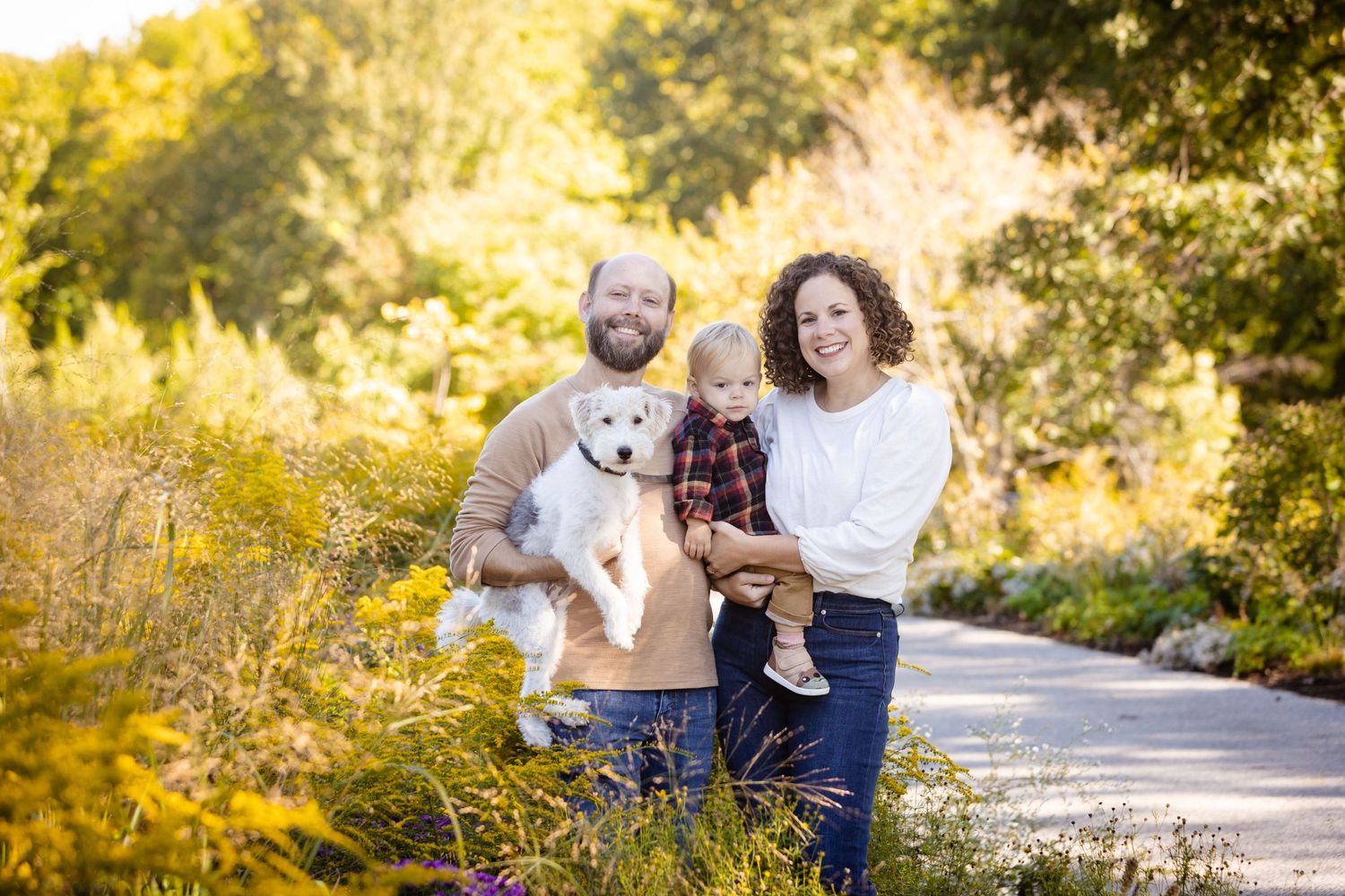 A couple and their fluffy white dog enjoy a sunny autumn day on a walking path surrounded by golden foliage.