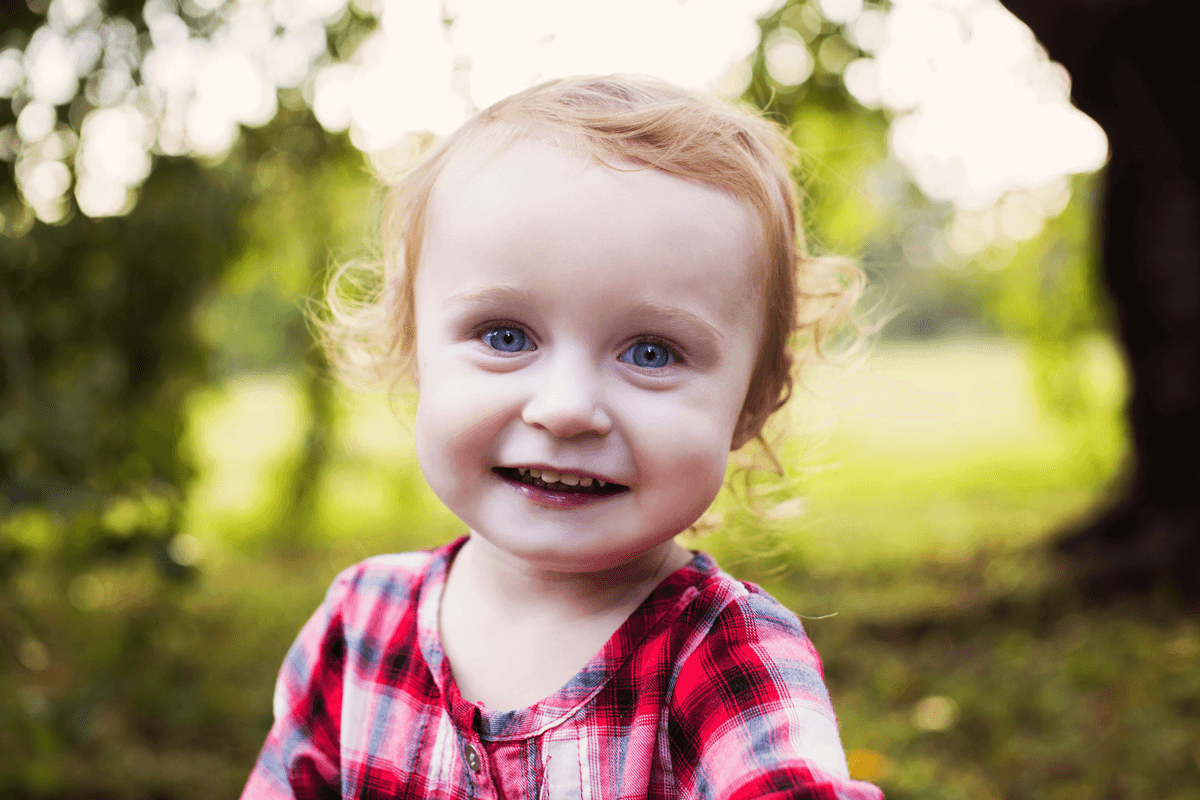 A cheerful toddler in a red plaid shirt smiles brightly outdoors.