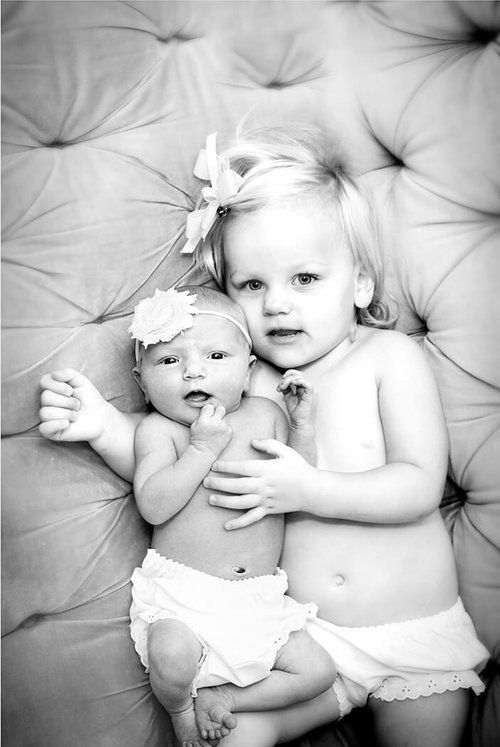 Two small children in white outfits laying on tufted grey surface in black and white photo.