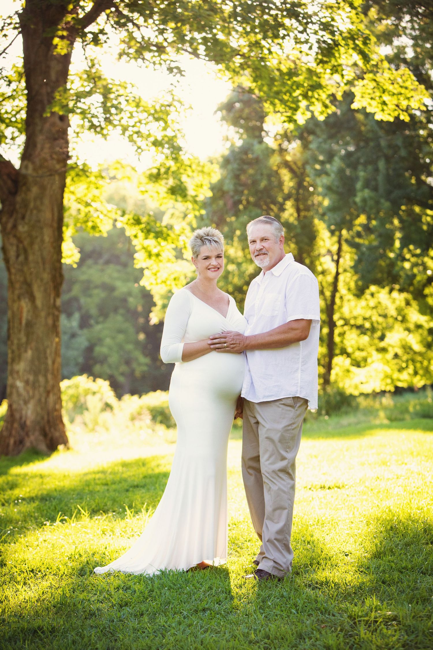 Expecting couple stands in warm sunlit outdoor setting with lush green trees and dappled golden light.