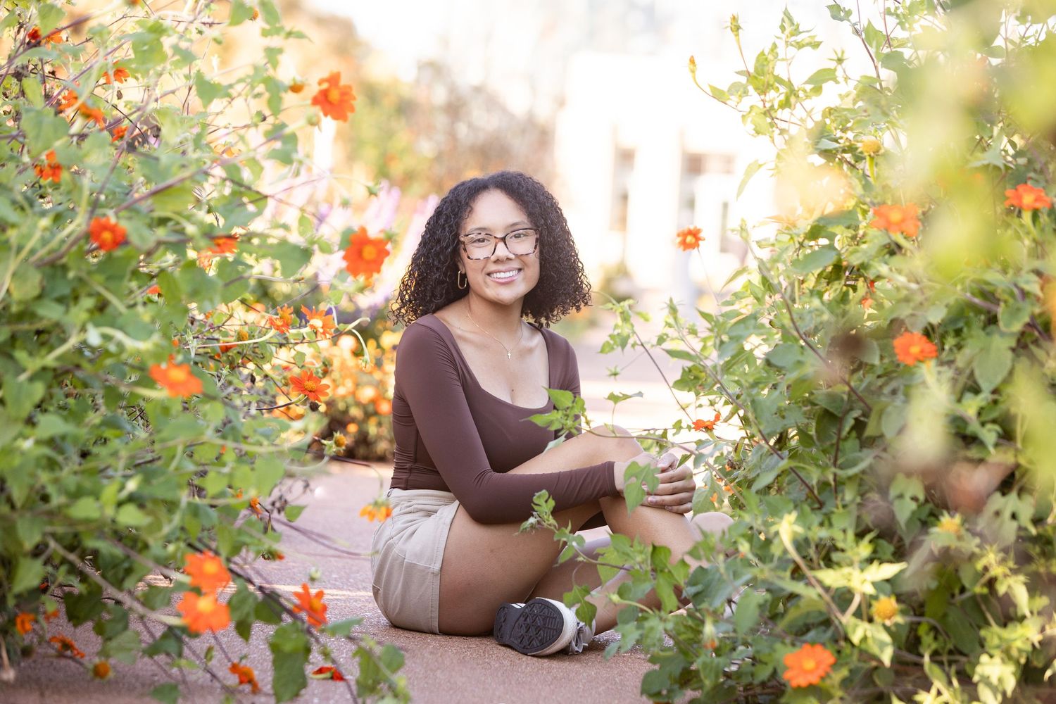 Young woman with glasses sitting cross-legged among vibrant orange flowers in a sunny garden, smiling warmly.